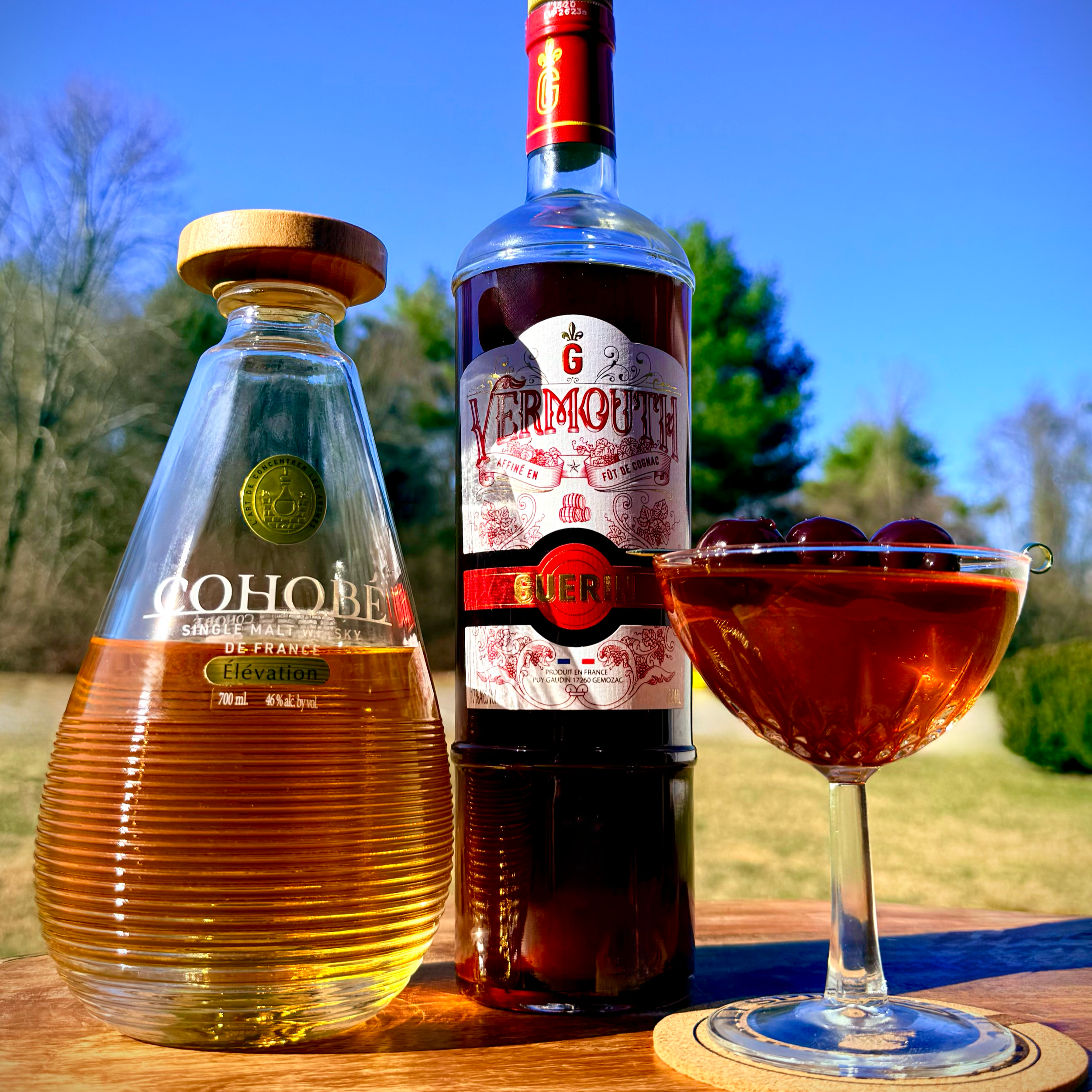 A teardrop bottle of Cohobé Whisky Élévation and a bottle of Guerin Sweet Red Vermouth stand behind a crystal coupe glass filled with an amber cocktail and garnished with three skewered Bada Bing cherries.