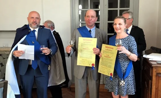Armagnac Musketeer in cape and blue sash presents new inductees Christine and Dan Cooney with blue sashes, medals, and certificates