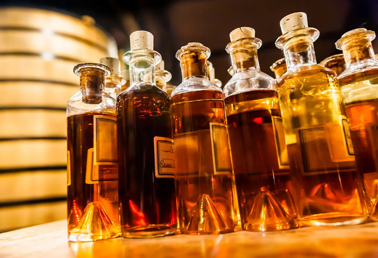 Collection of cork-toped samples of cognac with large barrel in the background, honeyed lighting