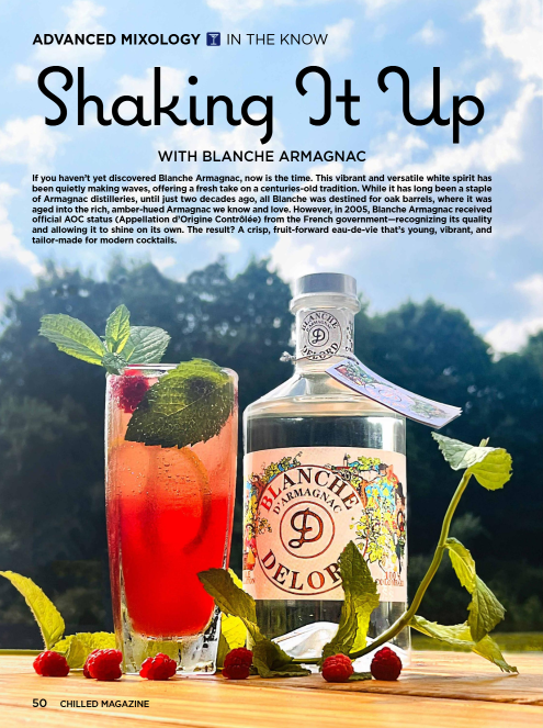 Close up on a reddish cocktail in a collins glass with ice, fresh, mint and raspberries next to a squat bottle of Blanche Armagnac Delord with trees and cloudy blue sky in the background and mint sprigs and raspberries in the foreground.
