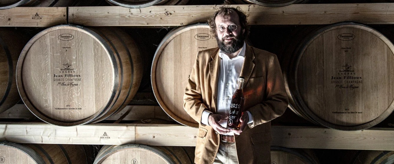 Bearded man with glassed in a camel coat and shirt holding a cognac tulip glass in front of shelves of cognac barrels.