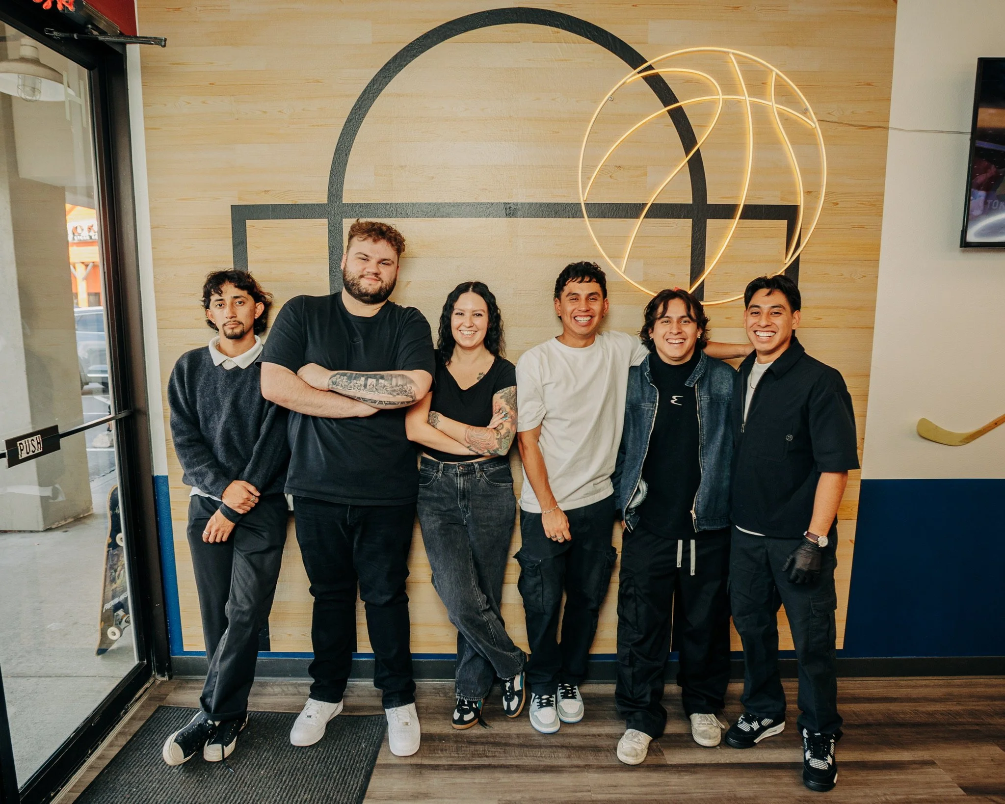 Group of six diverse young adults standing together inside a modern building, smiling at the camera, with a neon light art piece on the wall behind them.