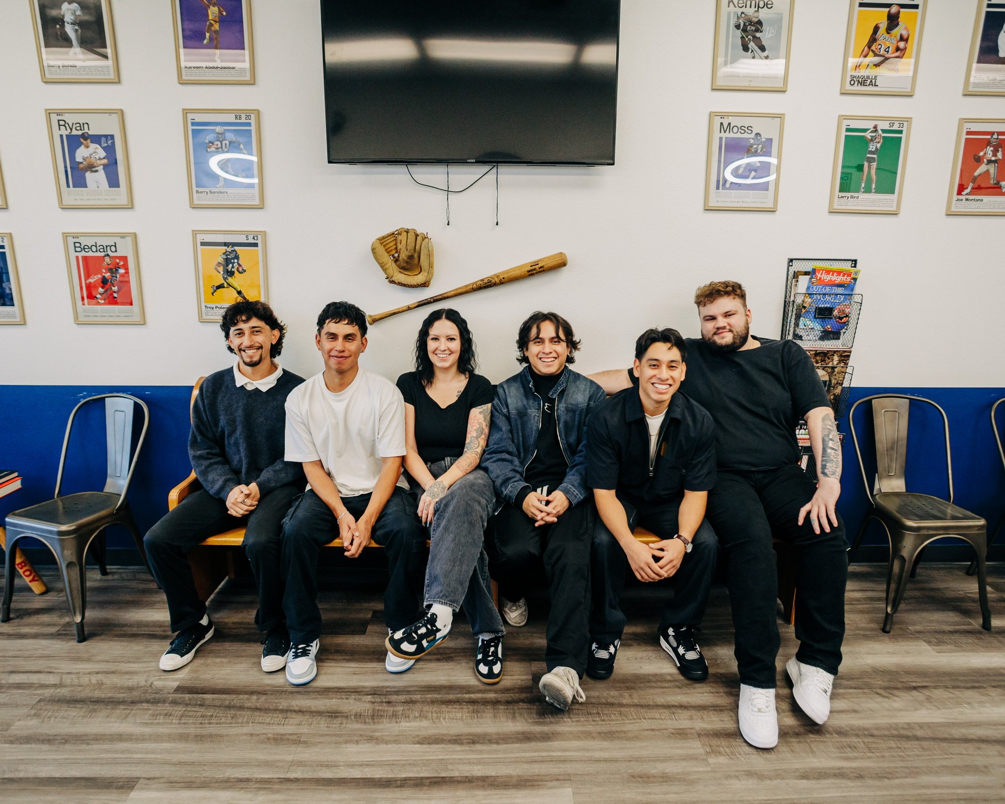 A group of six young adults sitting closely together on a bench, smiling and posing for a photo in an indoor space with framed sports posters on the wall and a large TV screen above them.