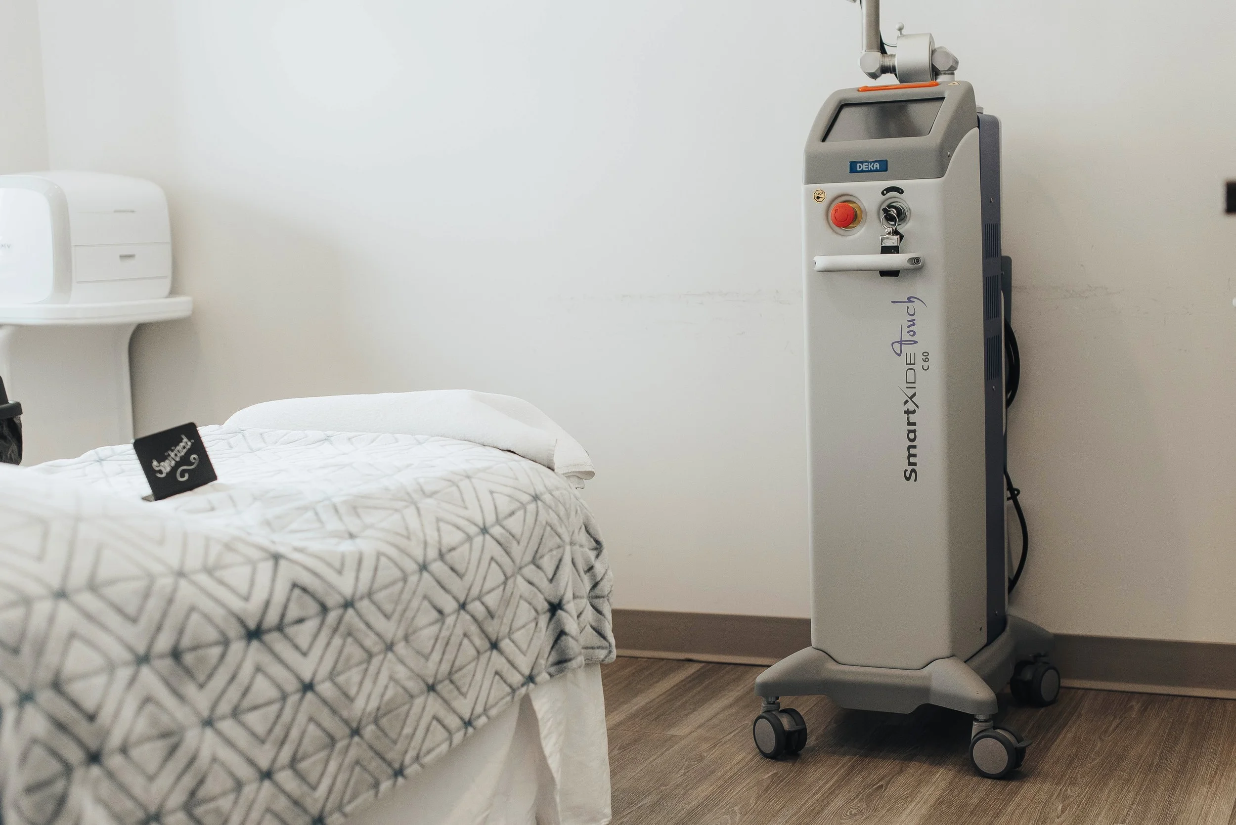 Medical room with a bed covered by a patterned blanket, a small table with medical equipment, and a tall medical device on wheels beside the bed.