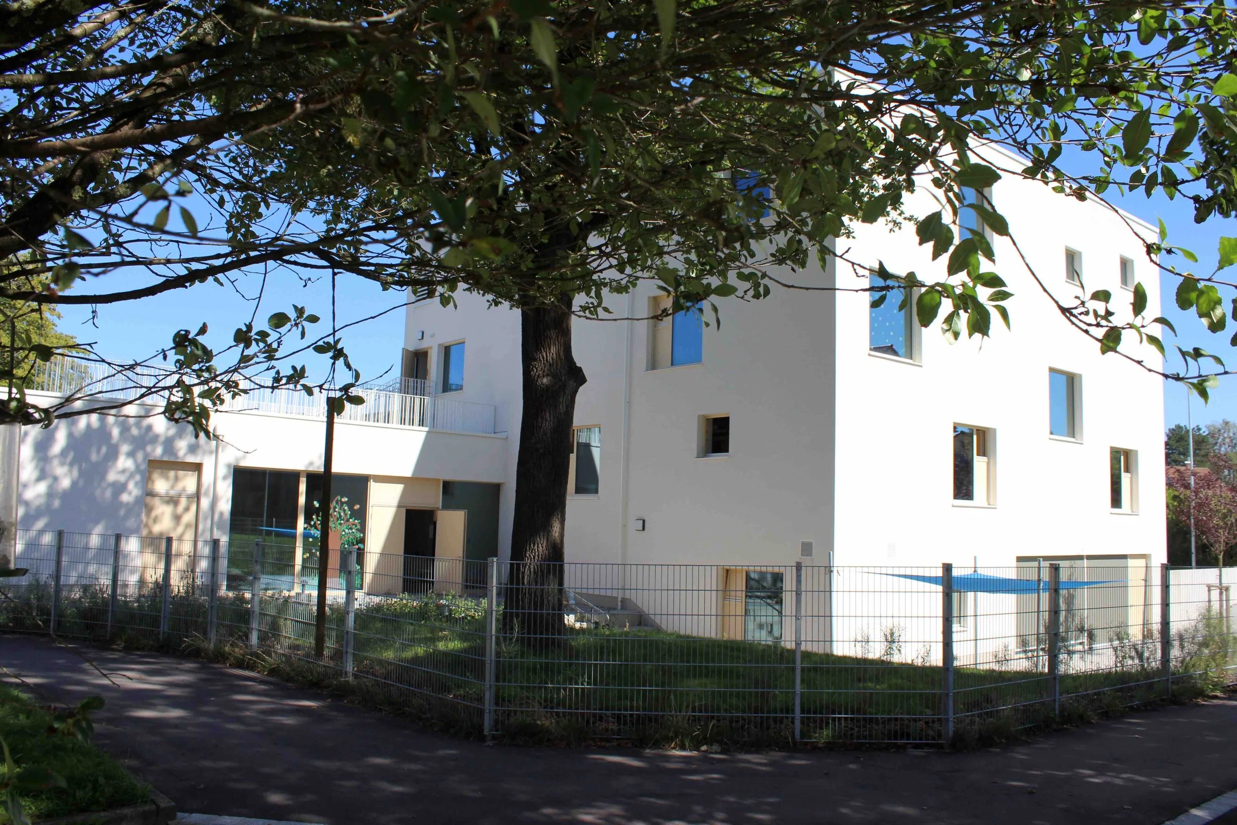 Weißes mehrstöckiges Gebäude mit mehreren Fenstern, umgeben von einem Garten und Zaun, im Schatten eines großen Baumes, blauer Himmel im Hintergrund.