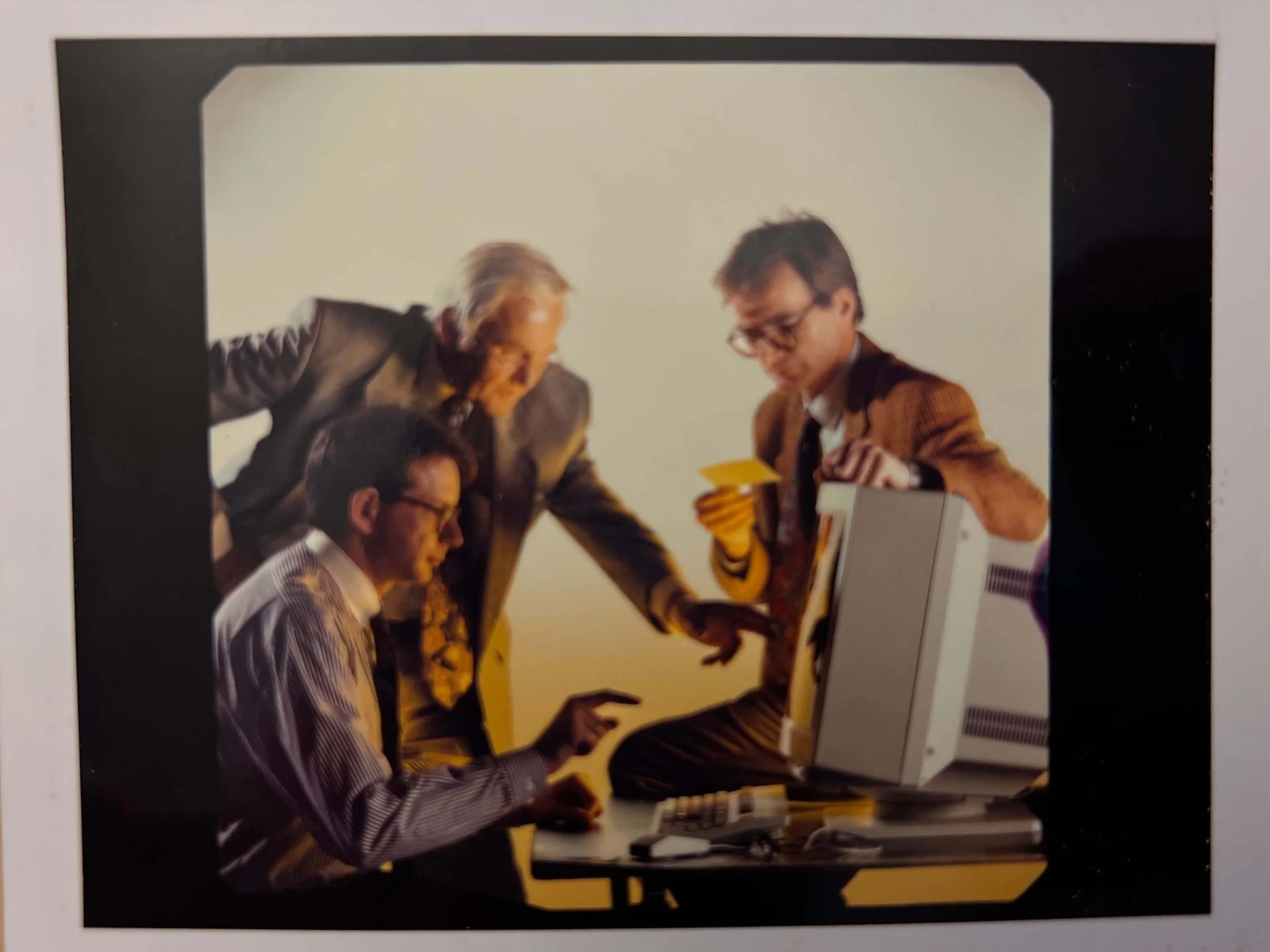 Drei Männer im Büro, einer sitzt vor einem Computer, der andere steht und zeigt auf den Bildschirm, während der dritte eine Karte hält.