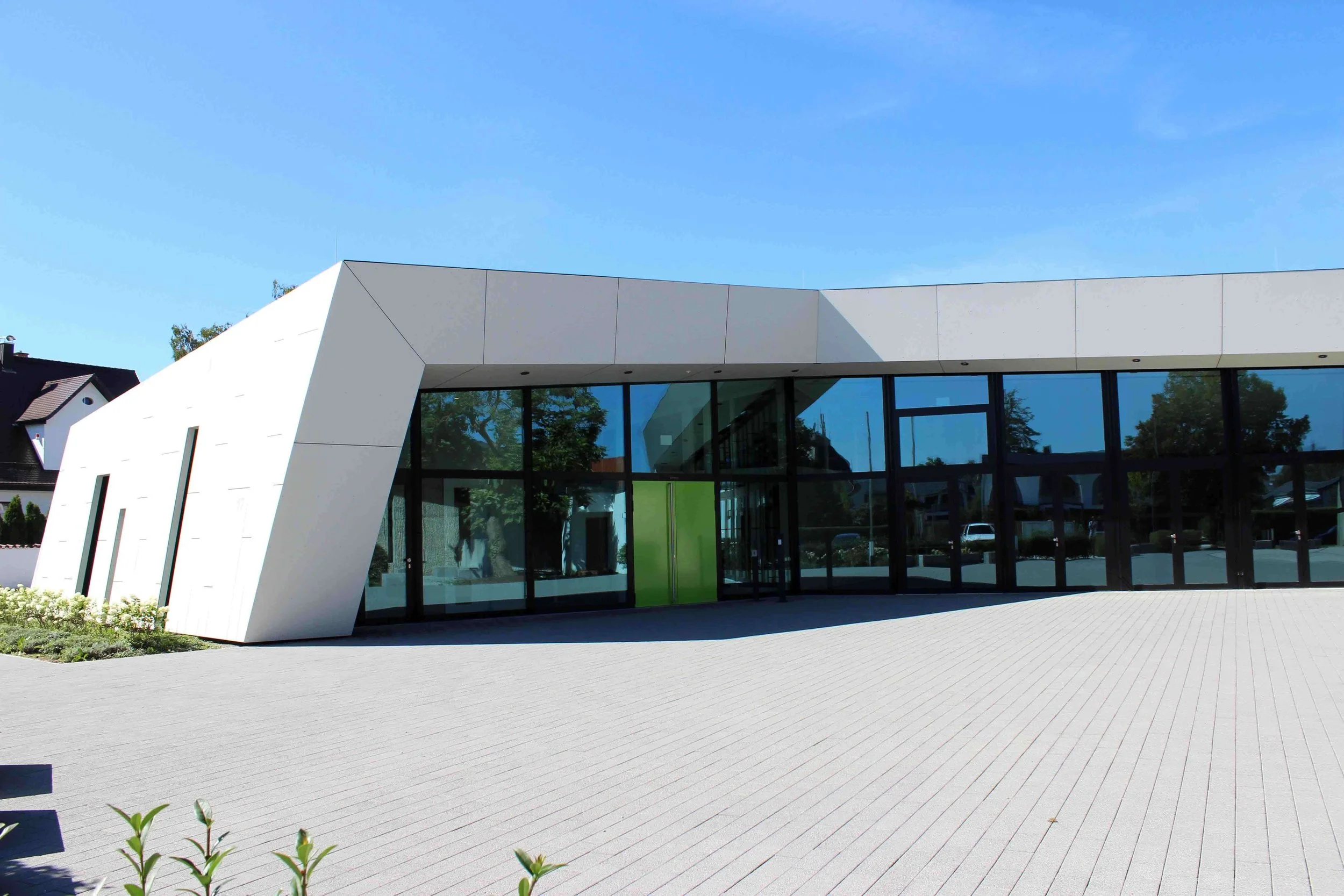 Modernes Gebäude mit großen GlasFenstern und weißen Wänden, umgeben von einem gepflasterten Platz.