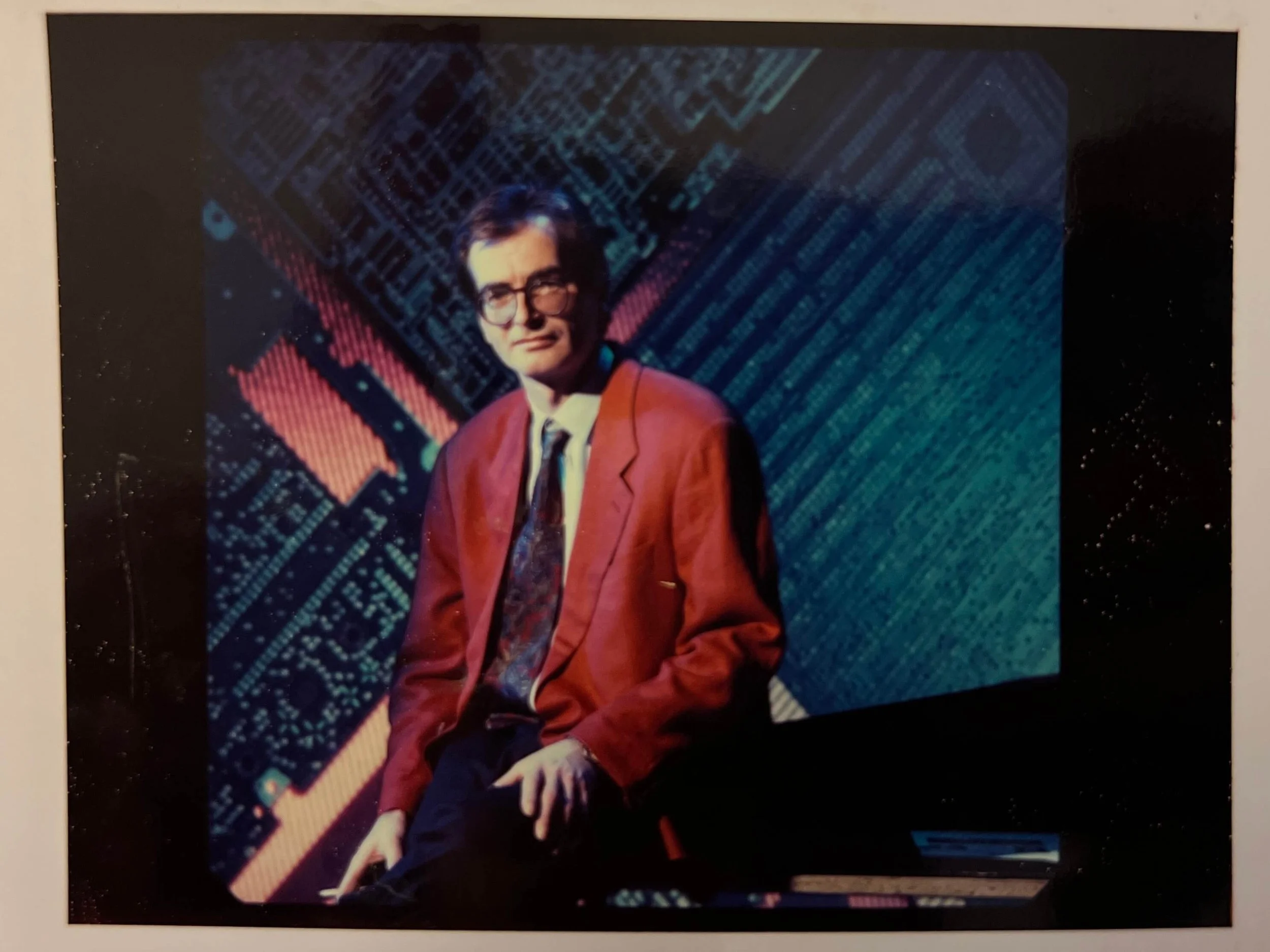 Ein Mann trägt eine rote Jacke, ein weißes Hemd und eine bunte Krawatte, er trägt Brille und sitzt vor einem digitalen Hintergrund mit blauen und roten Mustern.