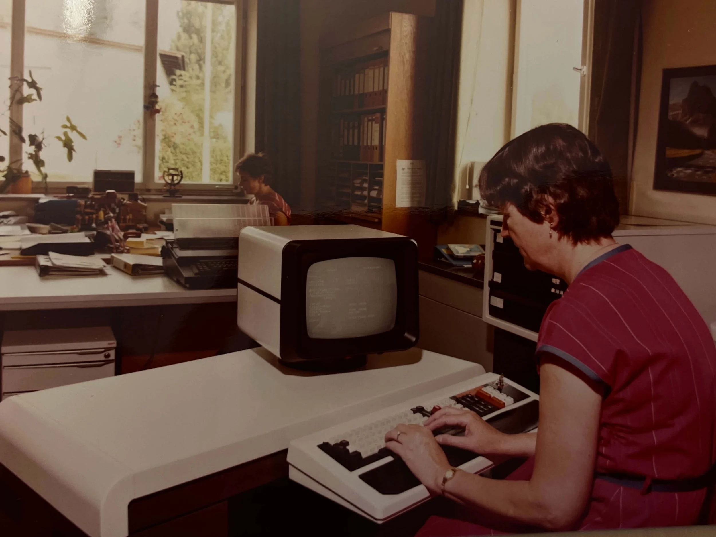 Eine Frau arbeitet an einem Computer in einem Büro, im Hintergrund ein Kind an einem Tisch.