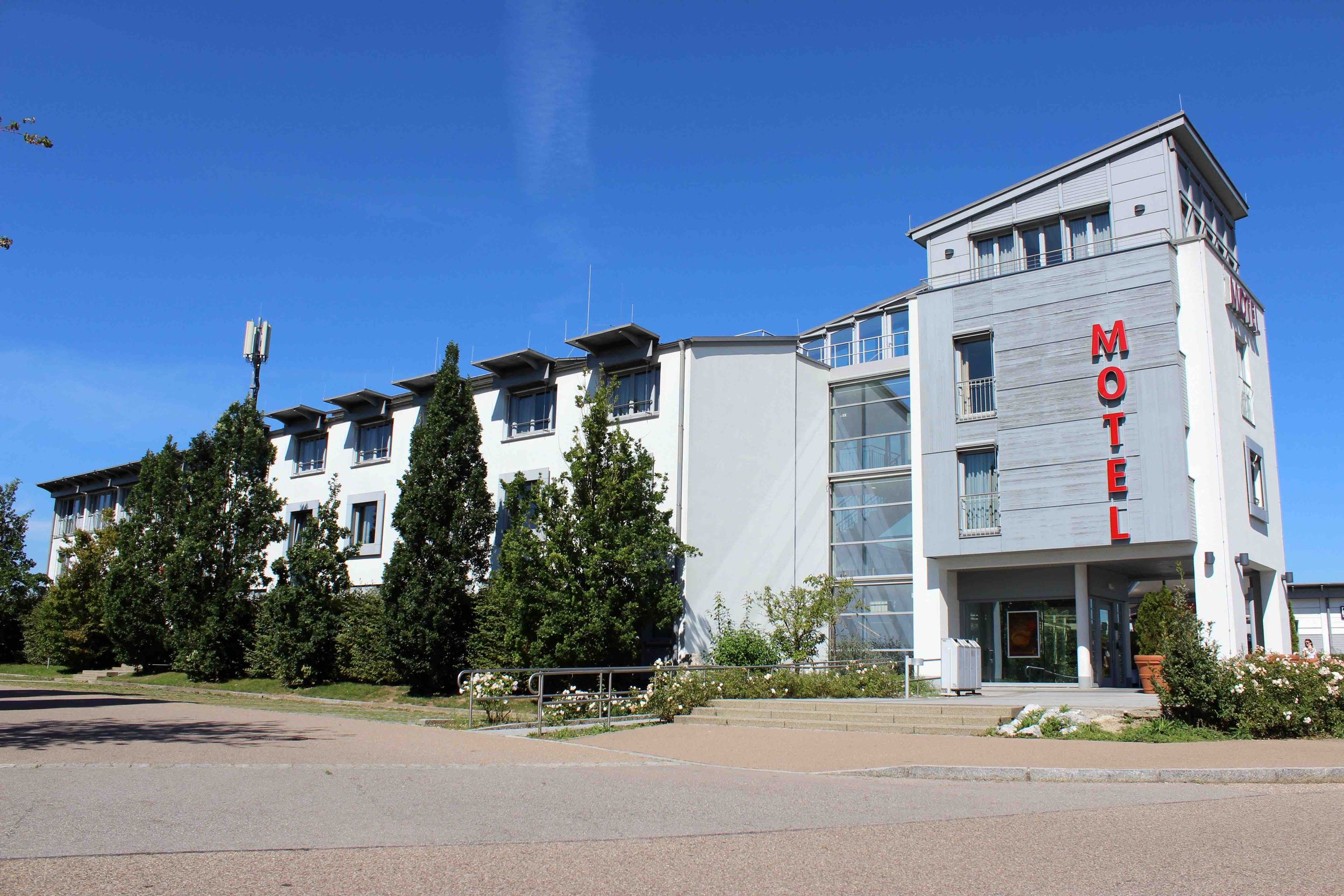 Modernes Hotelgebäude mit weißen Wänden, rotem Hotelschild, Bäumen im Vordergrund und einer blauen Himmelshintergrund.