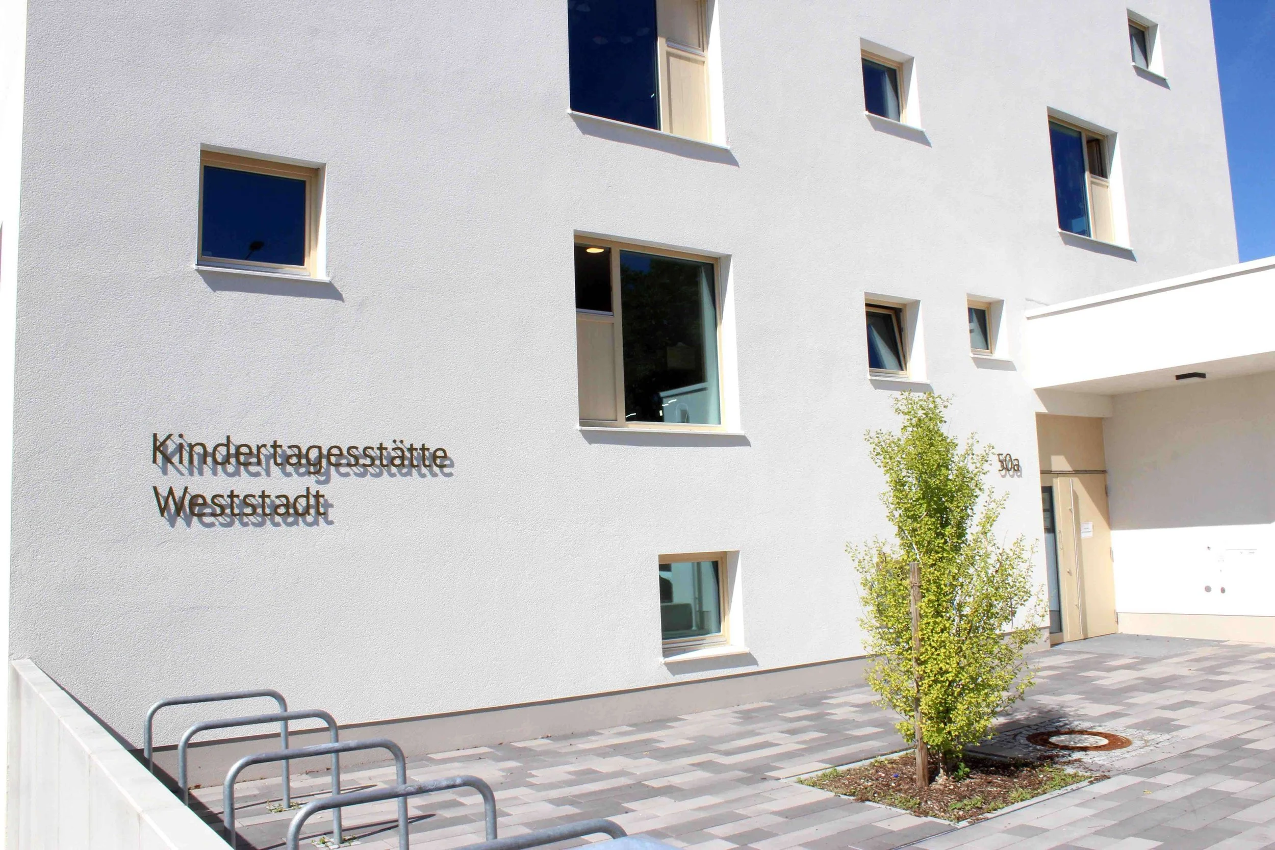 Das Foto zeigt das weiße Äußere eines Gebäudes mit mehreren quadratischen Fenstern, einer Tür und einem kleinen Baum vor dem Eingang. An der Wand ist ein Schild mit der Aufschrift "Kindertagesstätte Weststadt".