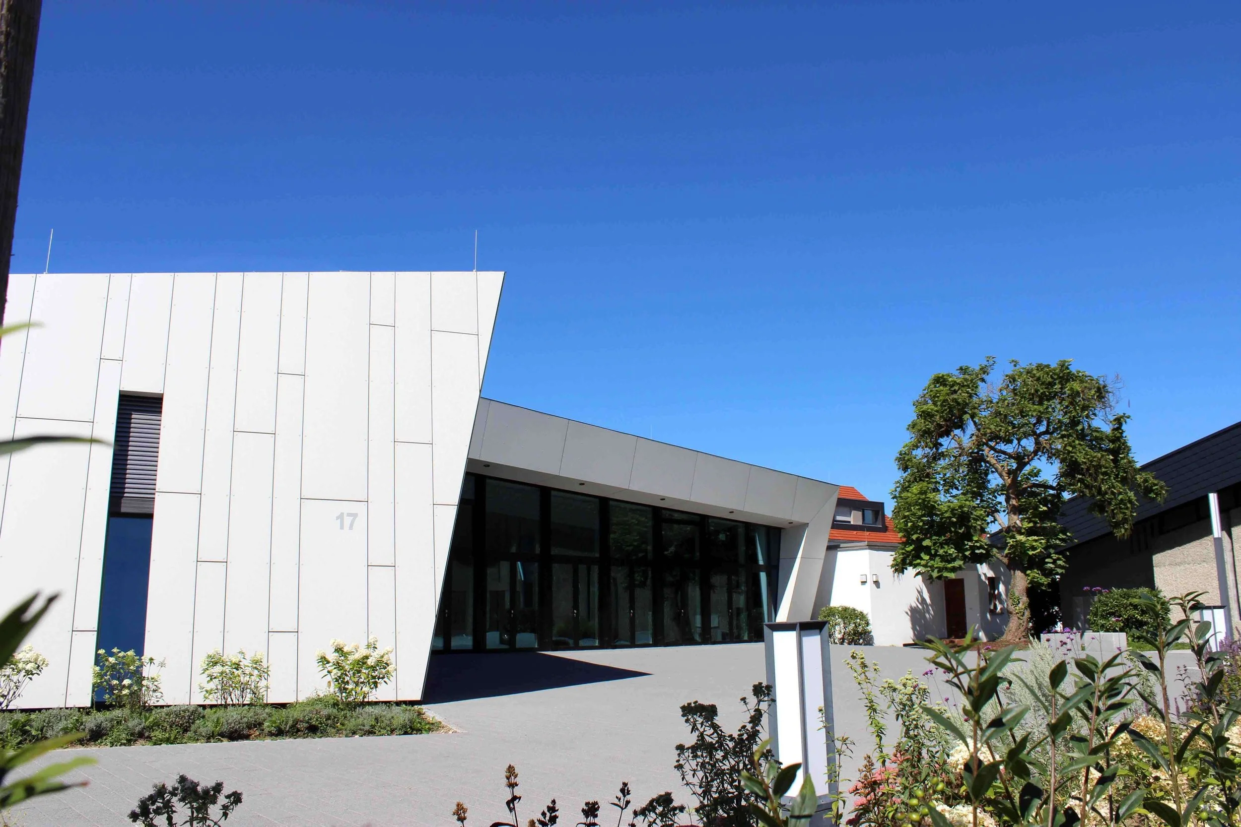Modernes, weißes Gebäude mit großen Glasfenstern, umgeben von Pflanzen und einem Baum, bei strahlendem blauen Himmel.