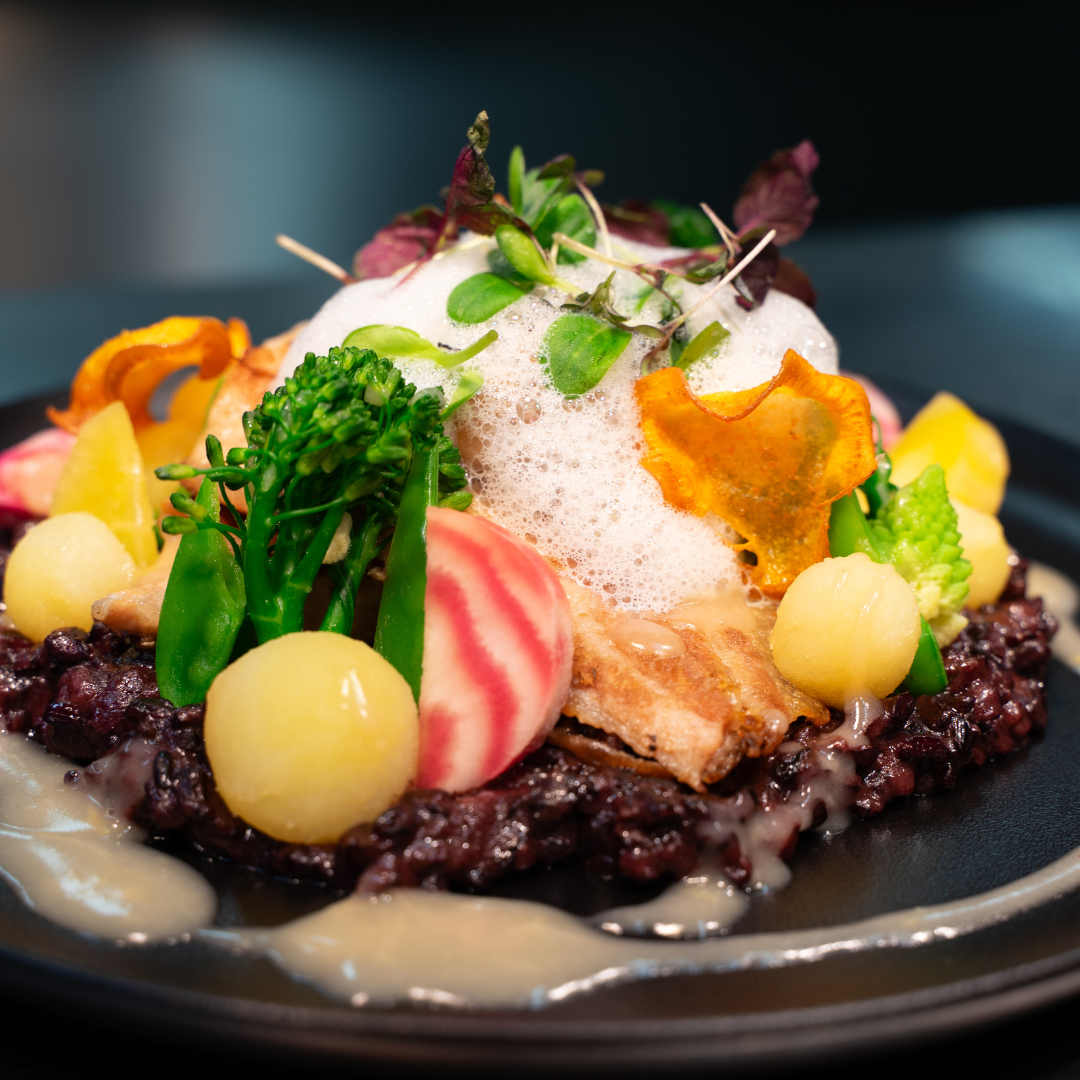 Ein Teller mit vegetarischer Mahlzeit, bestehend aus schwarzen Bohnen, buntem Gemüse, Kresse, Schaum und dekorativen Gemüseblumen.