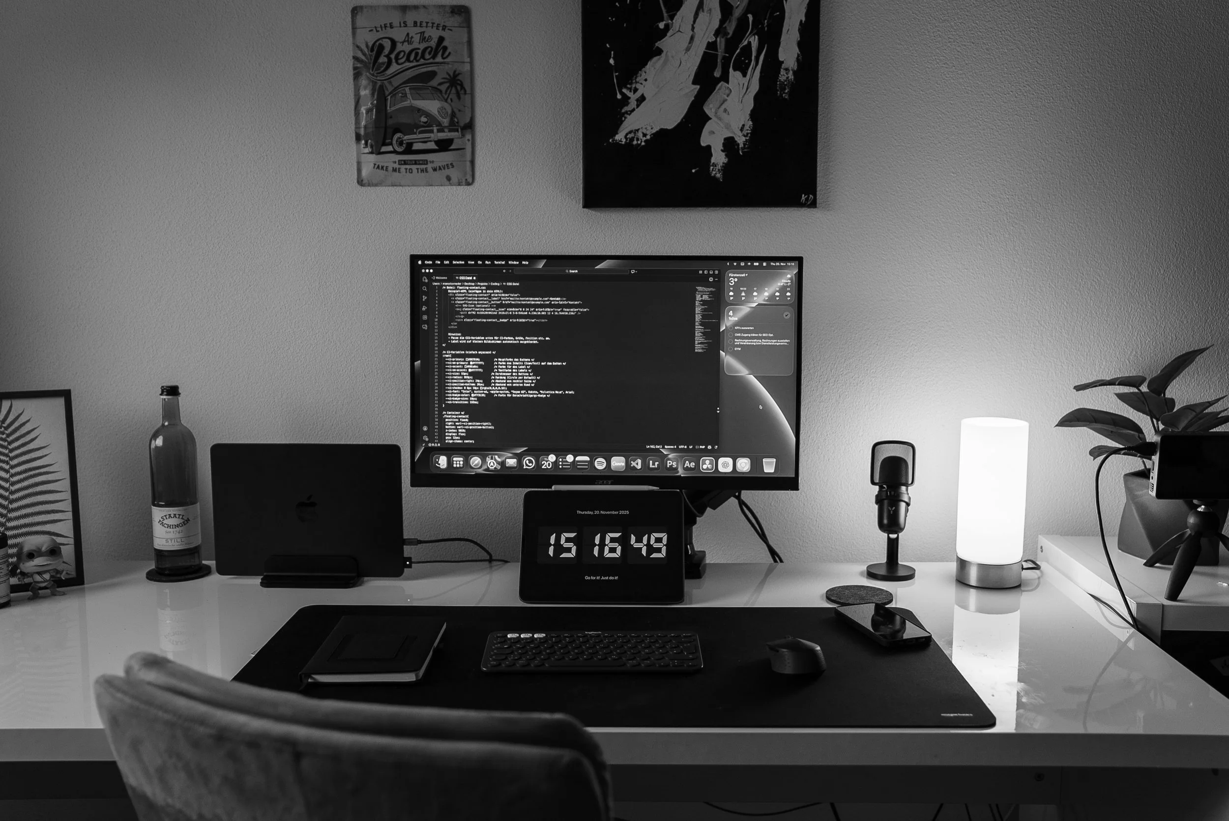 Ein Schreibtisch mit einem Computerbildschirm, einer Uhr, Tastatur, Maus, Smartphone, einem Notizbuch, einem kleinen Mikrofon, einer Lampe und Dekorationsgegenständen, darunter Pflanzen und Rahmen, in einem modernen Arbeitszimmer.