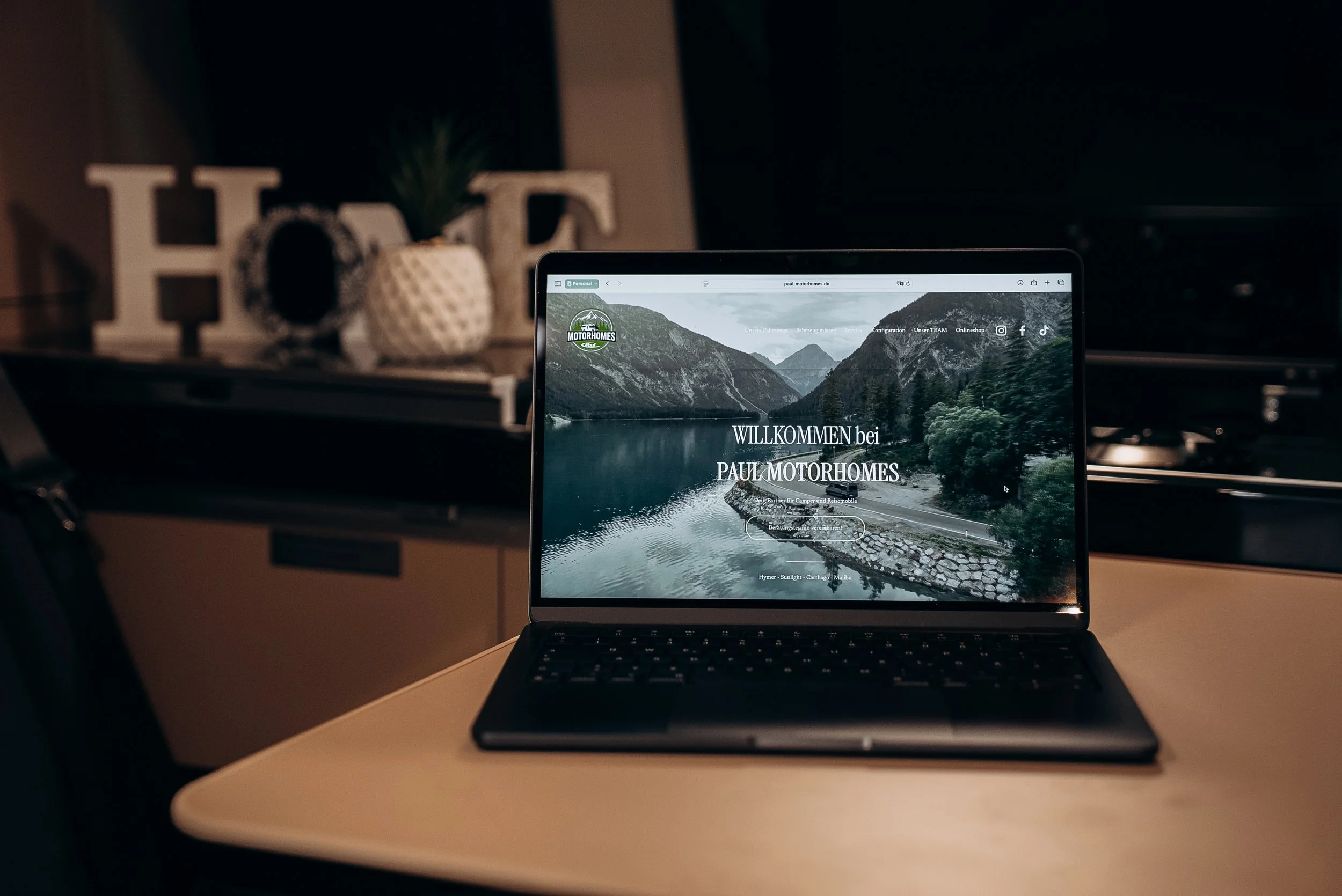 Laptop auf einem Tisch zeigt die Webseite von Paul Motorhomes mit einem Bild von einem See und Bergen im Hintergrund.