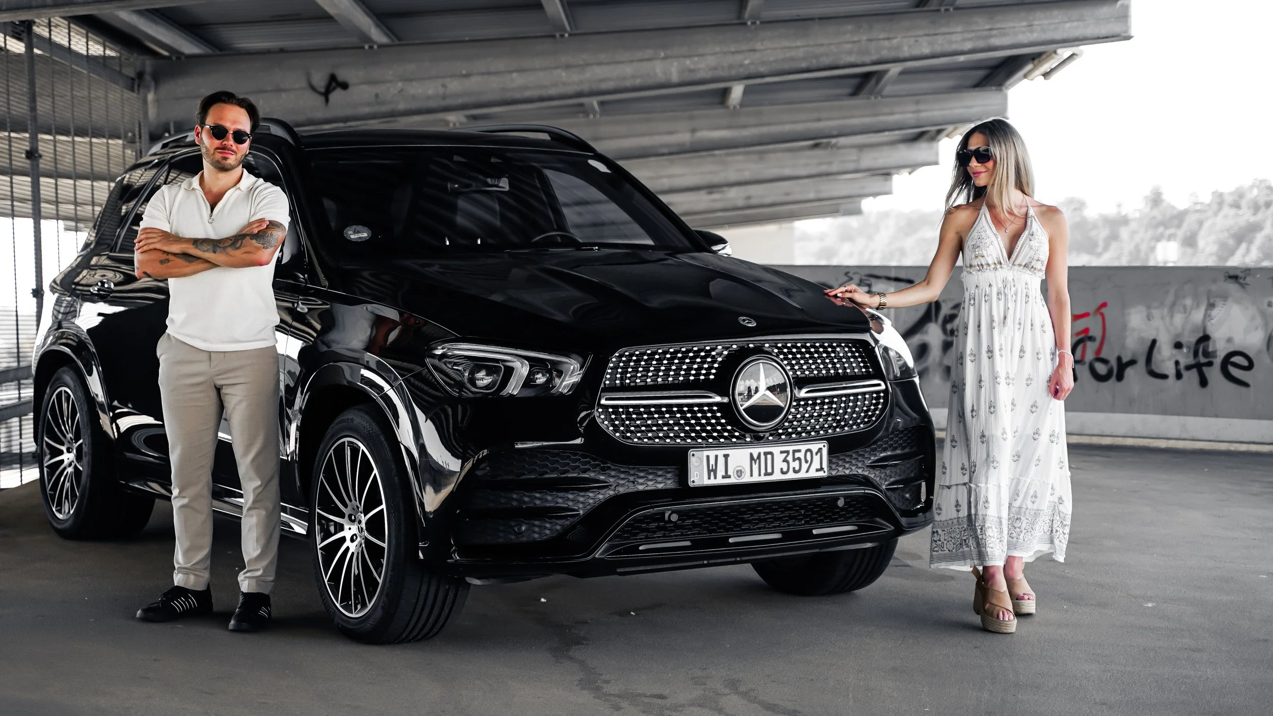 Ein Mann und eine Frau stehen neben einem schwarzen Mercedes SUV in einer Parkgarage. Der Mann trägt eine weiße Polo-Hemd, beige Hosen und dunkle Sonnenbrillen, die Frau trägt ein langes, weißes Kleid mit Mustern und Sonnenbrillen. Sie posieren in der Nähe des Autos.