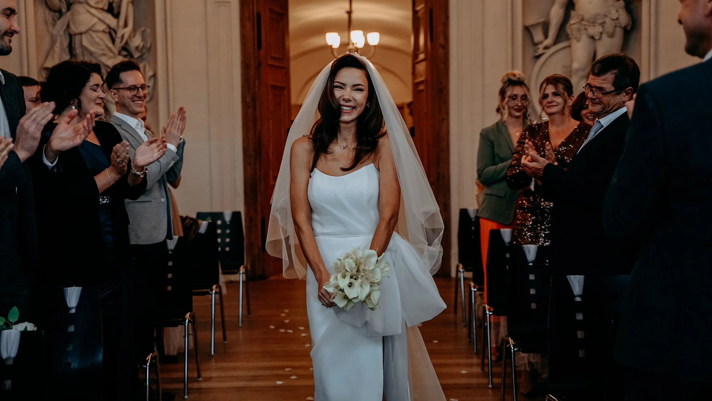 Braut in weißem Kleid mit Schleier hält Blumenstrauß und lacht während ihrer Hochzeit in einem festlich dekorierten Raum, umgeben von applaudierenden Gästen.