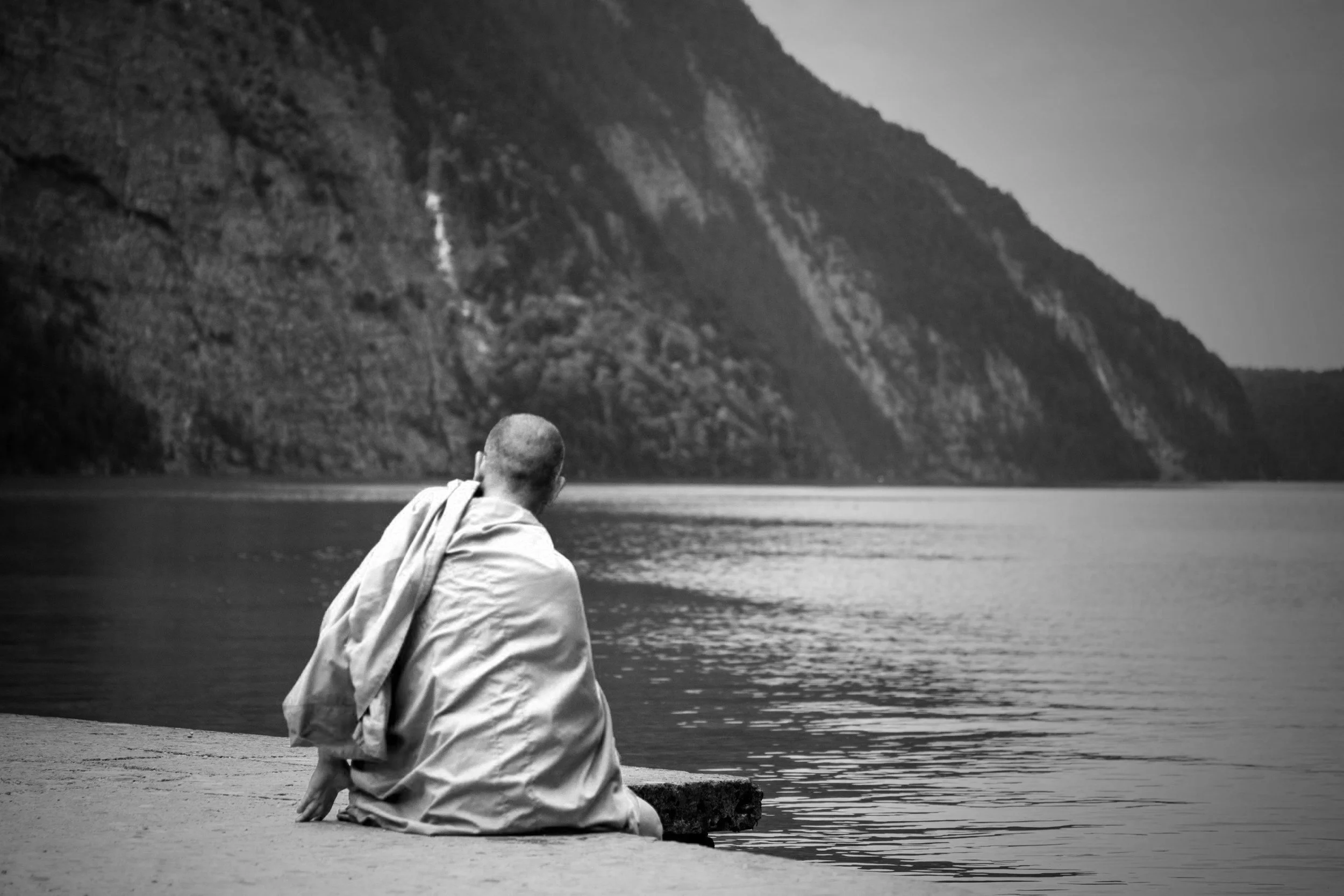 Ein Mann in einem Gewand sitzt am Flussufer und blickt auf das Wasser und die Berge im Hintergrund, Schwarz-Weiß-Fotografie.
