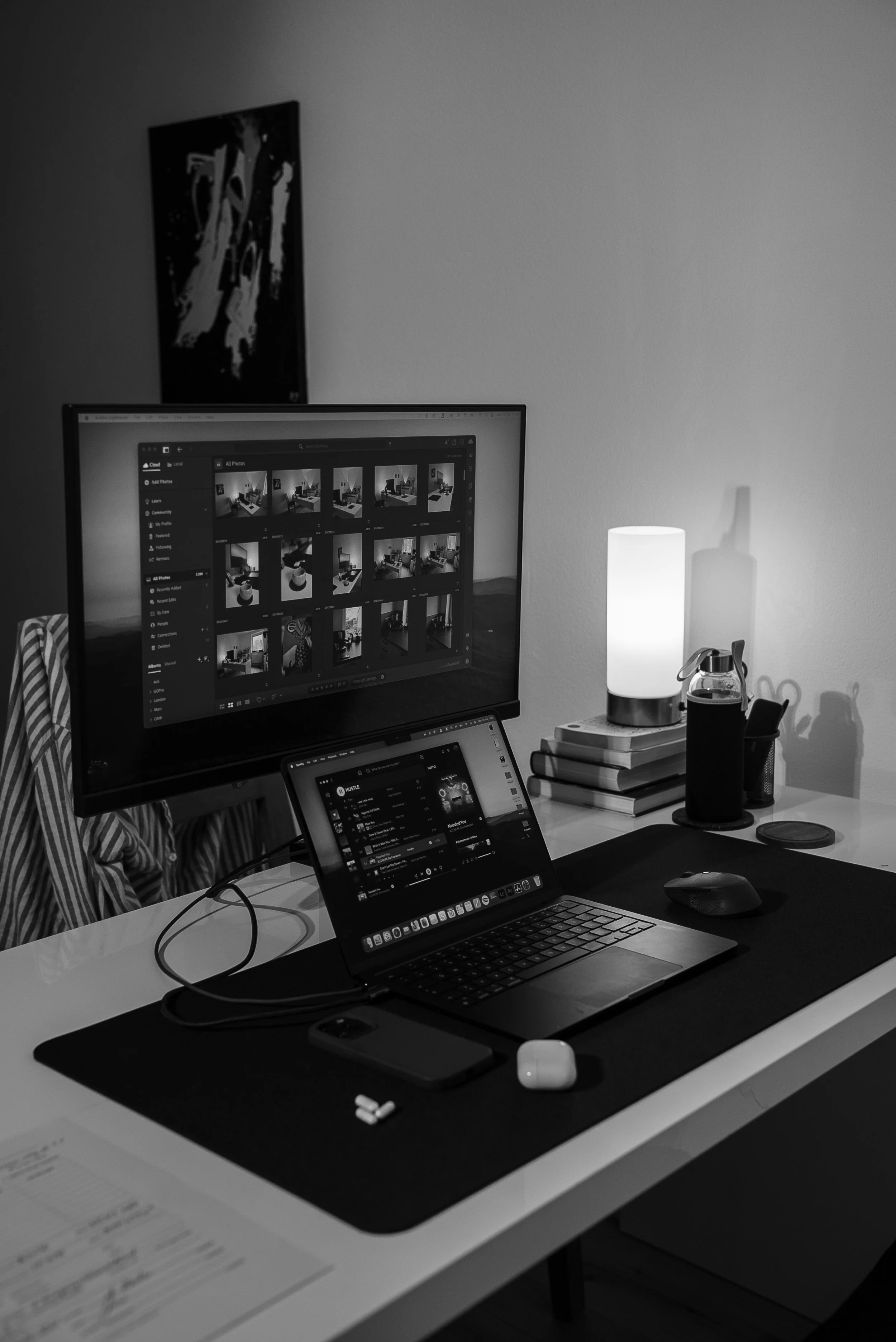 Schreibtisch mit Laptop, externem Monitor, Smartphone, Wireless-Kopfhörern, Wasserkocher, Büchern und Tischlampe in schwarz-weiß.