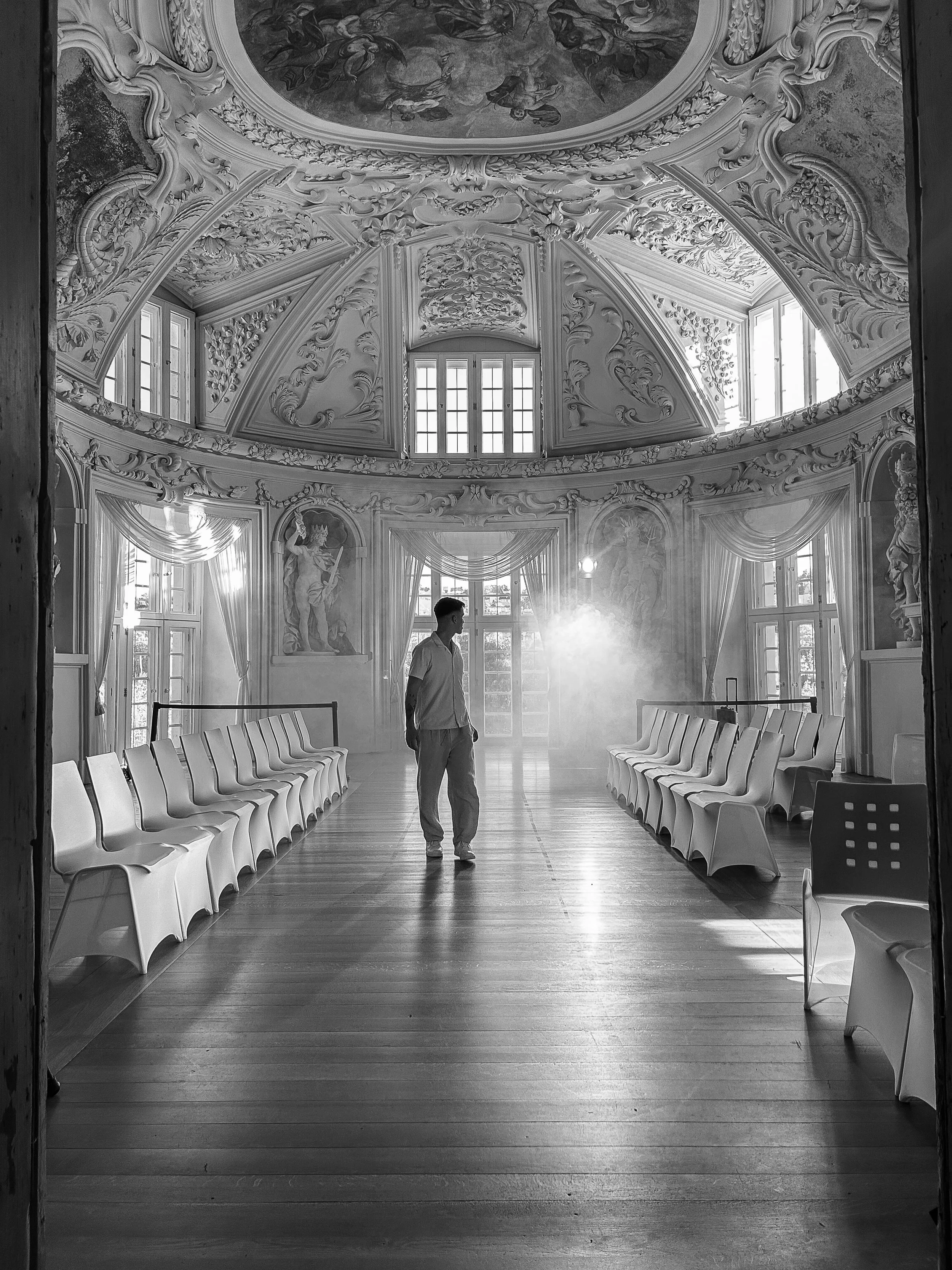 Ein eleganter Prunksaal mit barocker Decke und großen Fenstern, ein Mann steht in der Mitte des Raumes, umgeben von weißen Stühlen, halb im Schatten, im Hintergrund ist Licht, das durch die Fenster scheint.