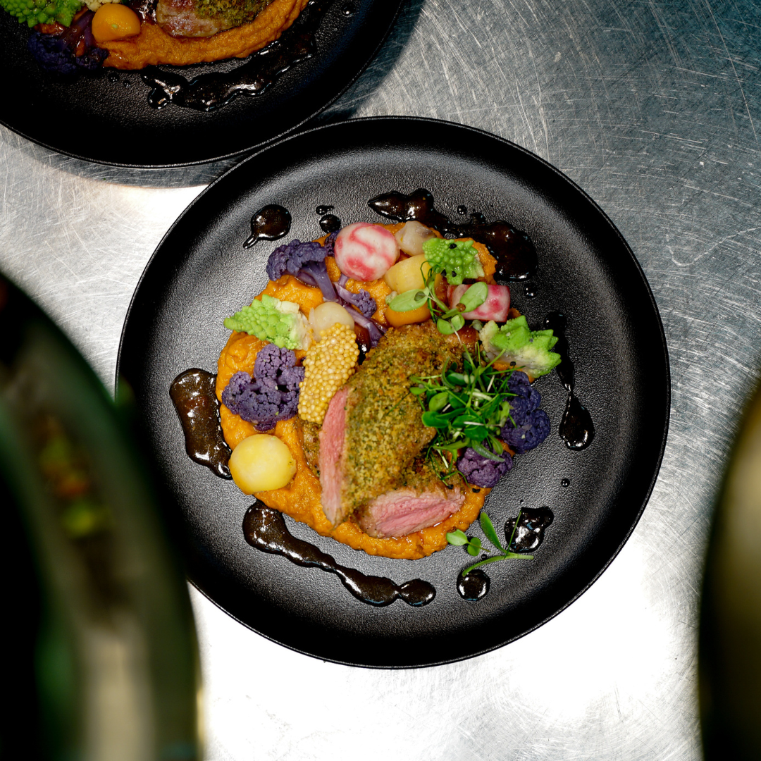 Gedünstetes Lammfilet mit Kräuterkruste, serviert auf einem Bett aus orangefarbener Gemüsepüree, begleitet von bunten Gemüsesorten und dunkler Balsamico-Reduktion, garniert mit frischen Microgreens auf einem schwarzen Teller.