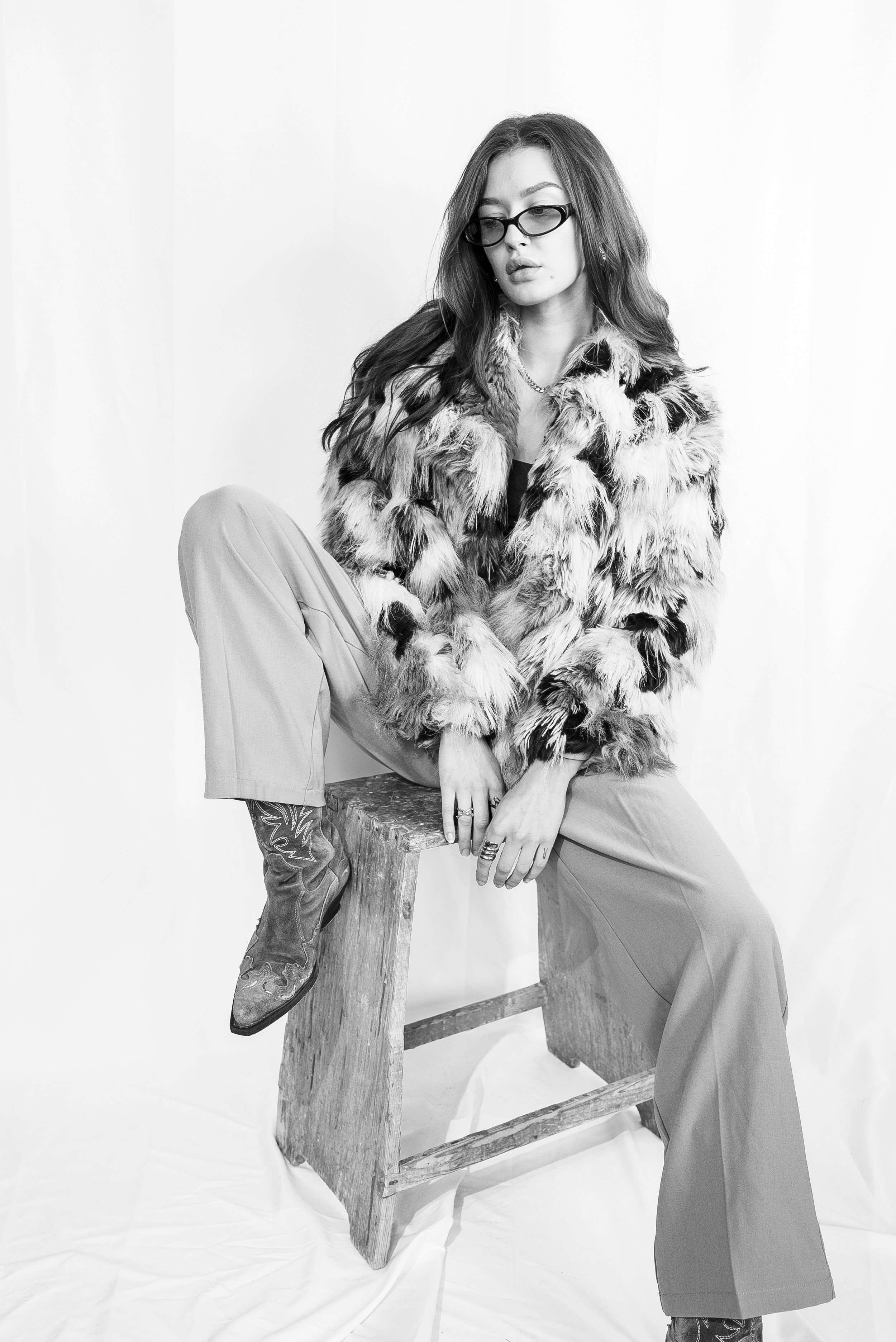 Schwarz-weiß-Foto einer jungen Frau mit langen, gewellten Haaren, Sonnenbrille und Pelzmantel, sitzend auf einem Holzhocker, im Studio vor einem weißen Hintergrund.