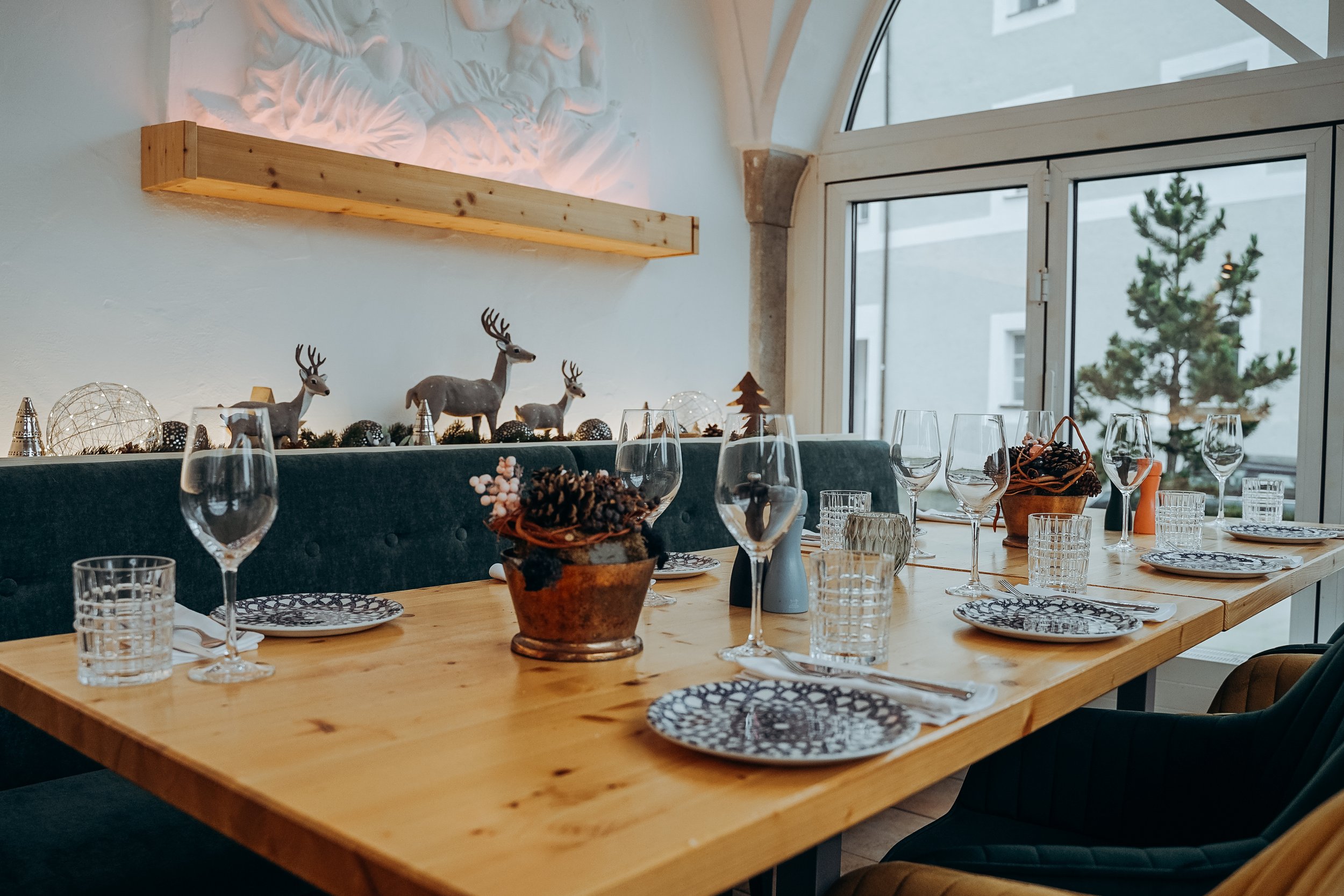 Ein festlich gedeckter Esstisch mit Wein- und Wassergläsern, Tellern und Besteck, dekoriert mit Weihnachtsdekorationen wie Rentieren, Tannenzweigen und kerzen. Im Hintergrund sind große Fenster mit Blick auf einen Weihnachtsbaum draußen.