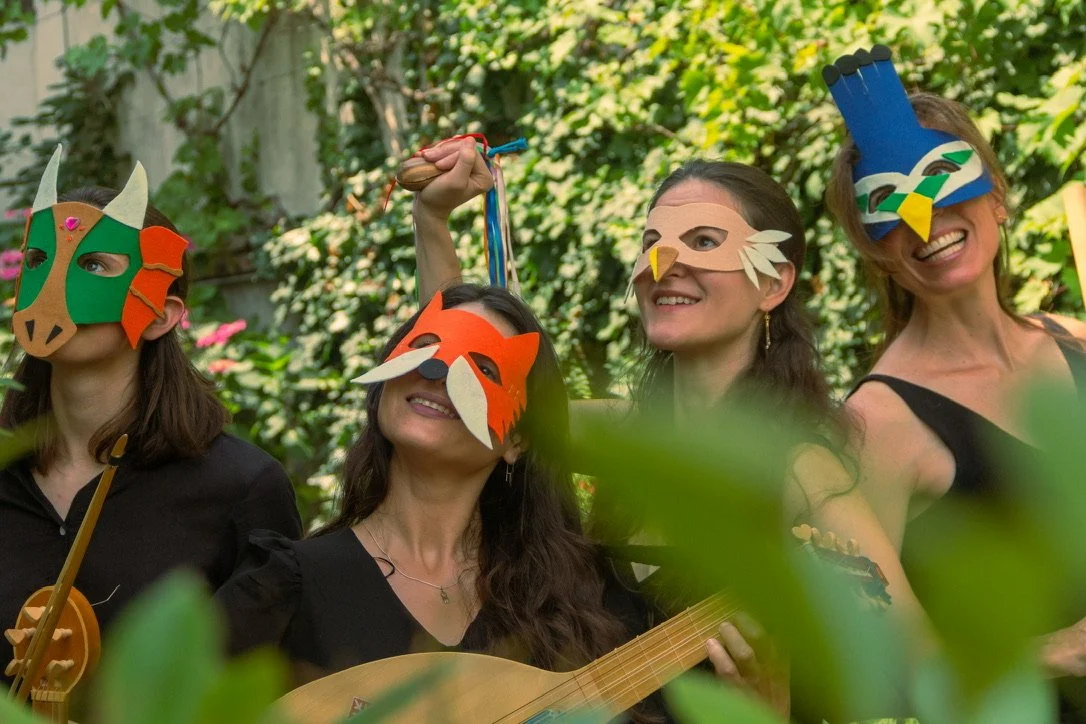 Four women wearing colorful animal masks, standing outdoors in a garden with green foliage and pink flowers, some holding musical instruments.