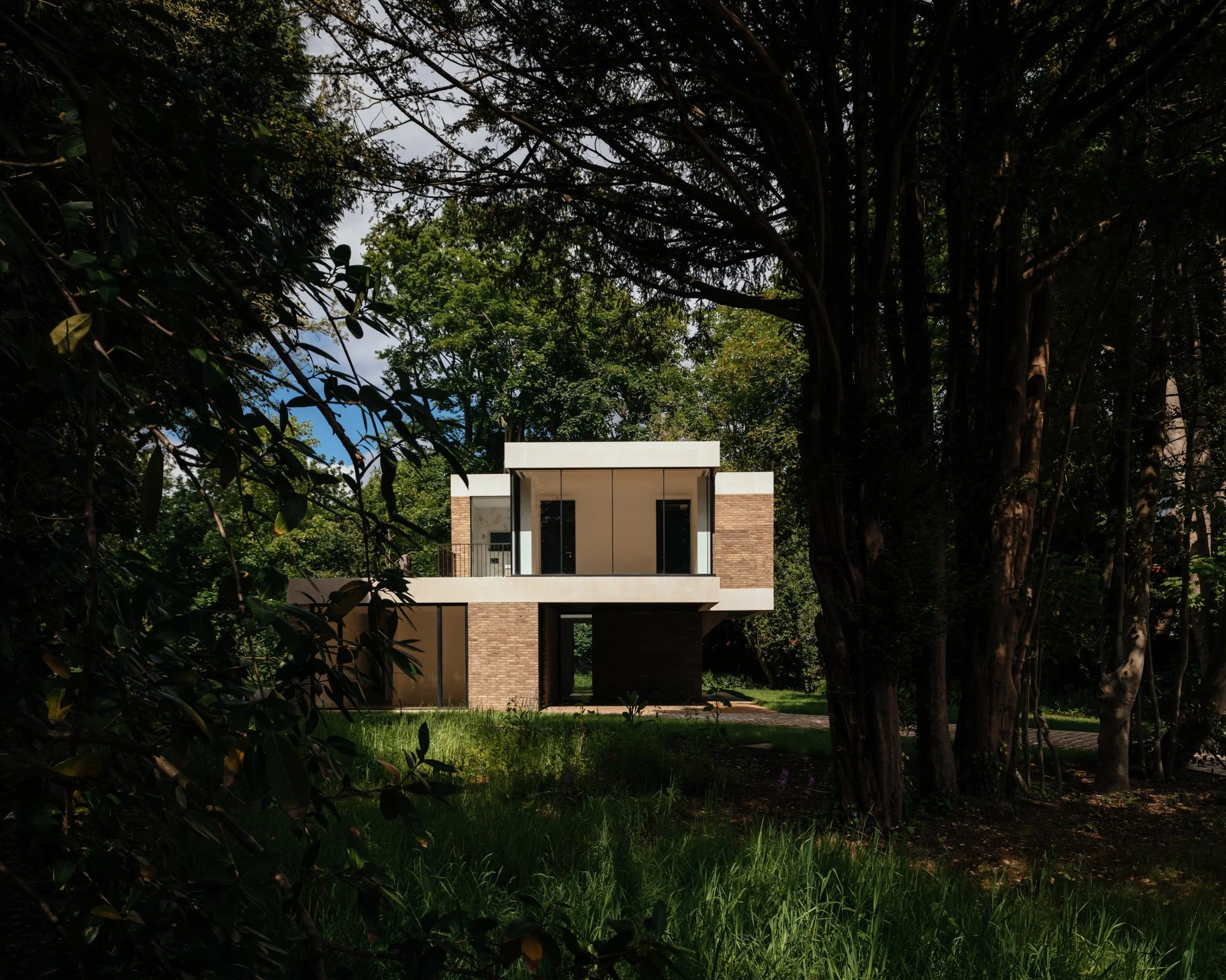Modern house with large glass windows and a flat roof, surrounded by tall trees and lush grass in a wooded area.