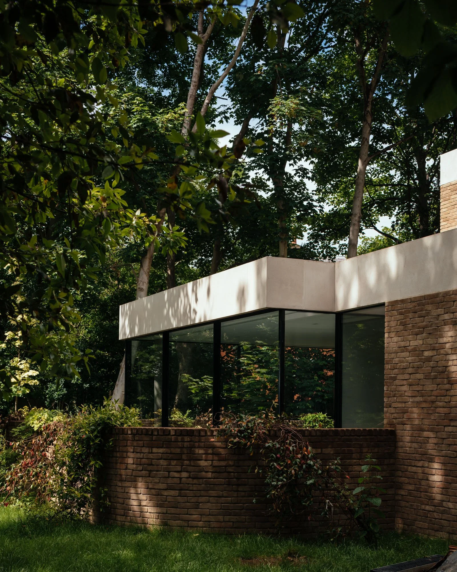 Modern house with large glass windows, white exterior walls, and a brick lower section, surrounded by lush green trees and plants