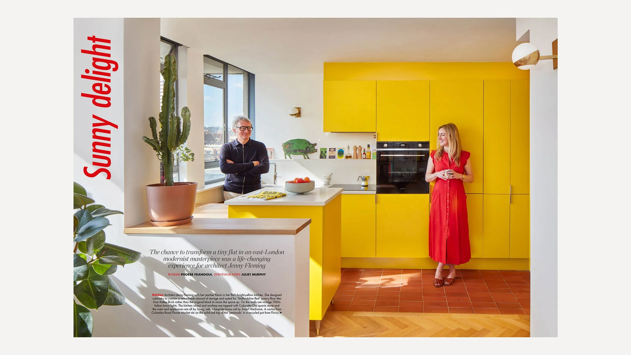 A bright yellow kitchen with large cabinets, a woman in a red dress holding a cup standing near the cabinets, a man with glasses and gray hair standing by the window, a cactus in a pink pot, and a bowl of oranges on the kitchen counter.