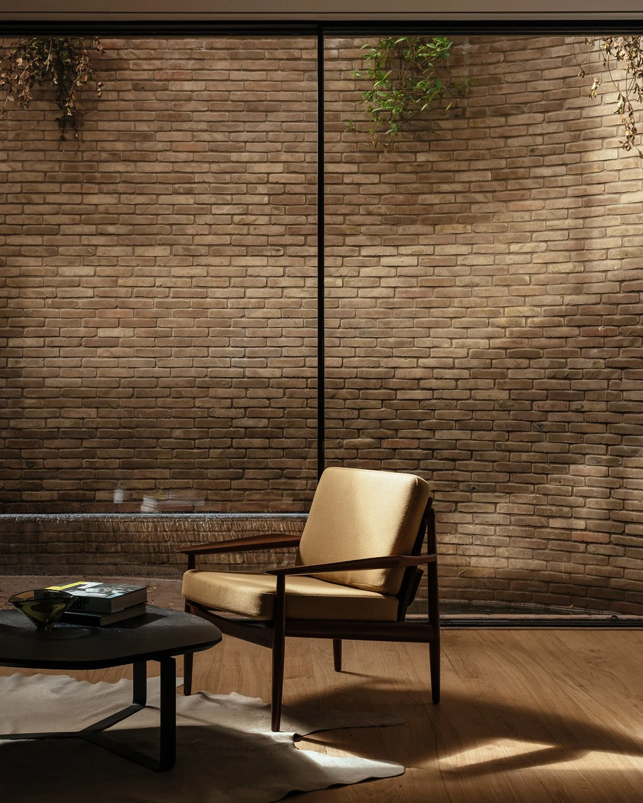 A cozy living room corner with a beige armchair, a black coffee table, and a brick wall in the background, with some greenery hanging from above.