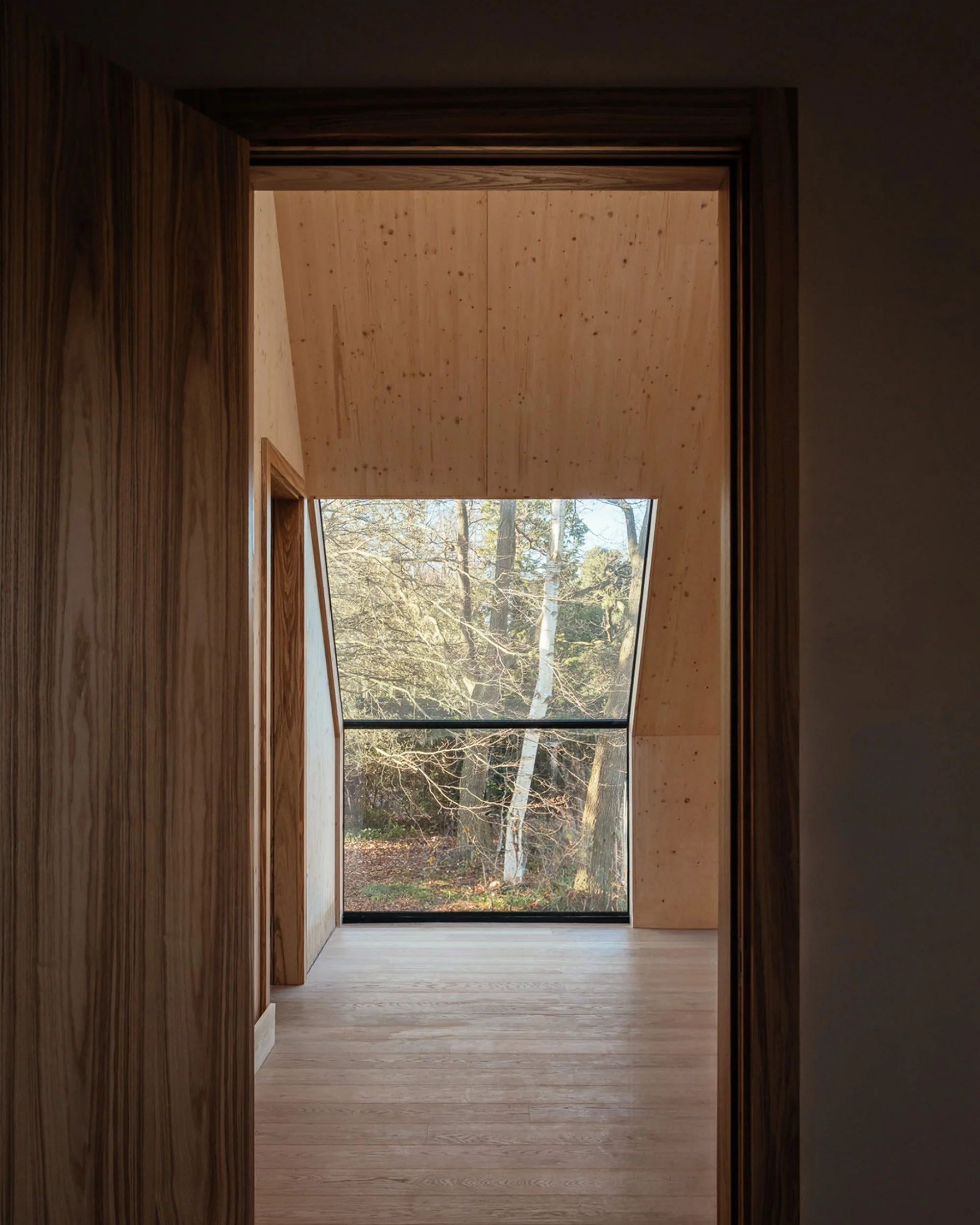 Interior view of a modern room with large window showing trees and sky, wooden walls and ceiling, and light wooden floor.