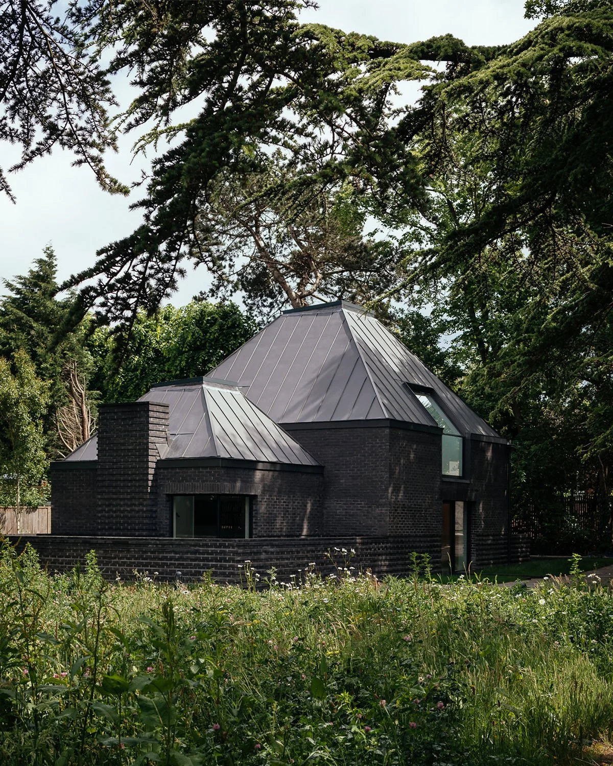 A modern house with black brick exterior and steep metal roof, surrounded by trees and greenery.