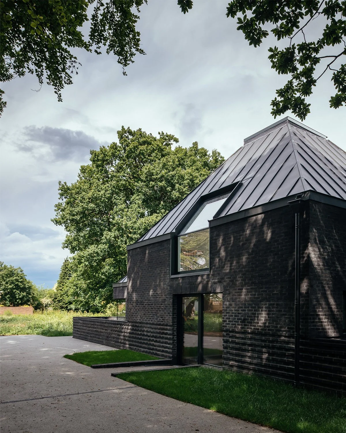 Modern black brick house with a steep metal roof, large windows, and surrounded by lush green trees.
