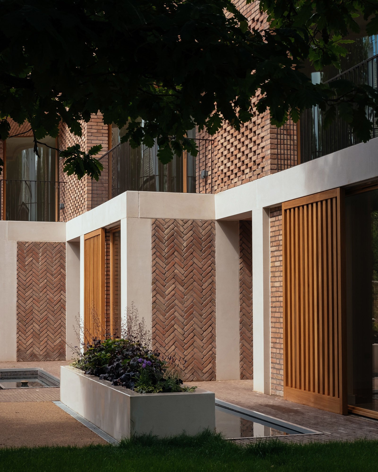 Modern building with brick and white concrete walls, wooden accents, a balcony with metal railing, a garden bed with flowers, and a water feature with a fountain in front.