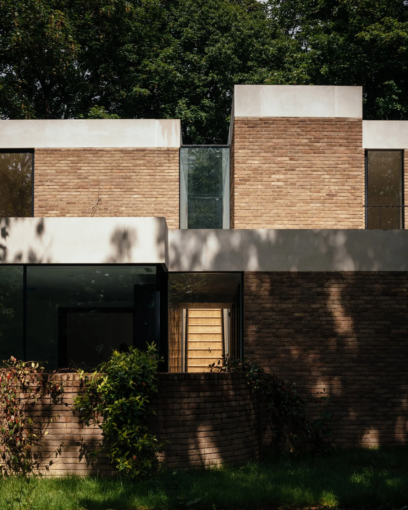 A modern house with brick walls and large windows, surrounded by greenery including trees and bushes, with sunlight casting shadows.