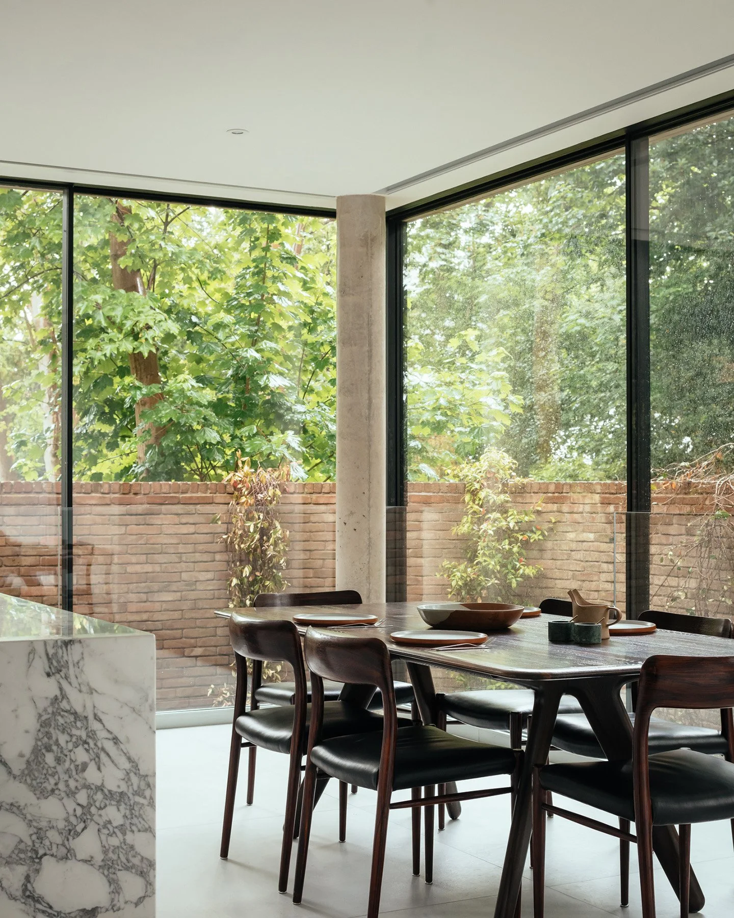 Modern dining area with a wooden table set with bowls, plates, and a teapot, surrounded by black chairs, large windows showing green trees, and a marble kitchen counter in the foreground.