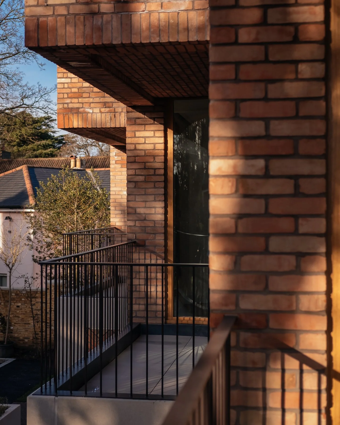 Close-up of a brick building with a glass door, black metal railing, and neighboring structures in the background, during daytime with sunlight casting shadows.
