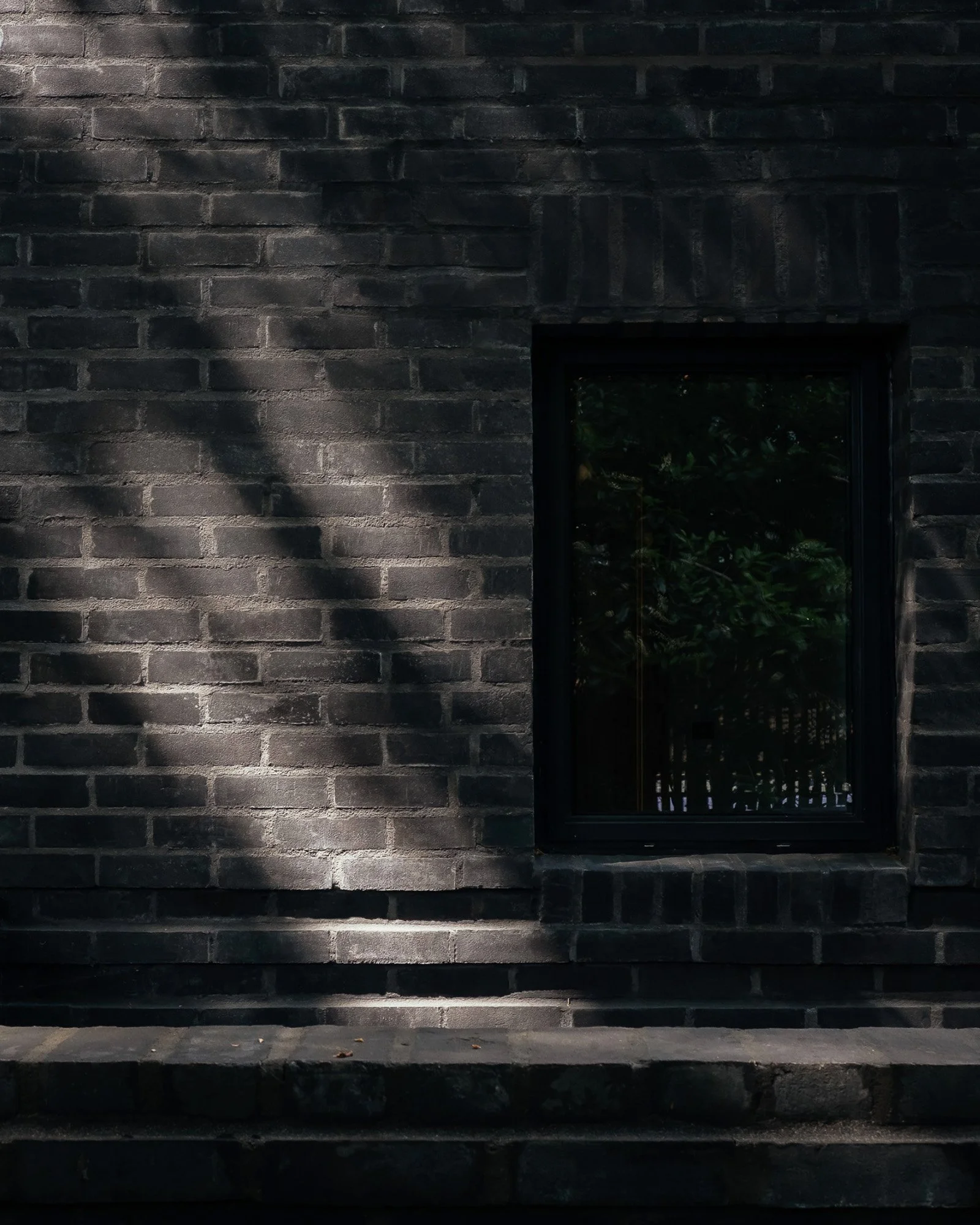 A dark brick wall with a small window and a shadow cast on the left side. A step and pavement are visible at the bottom.