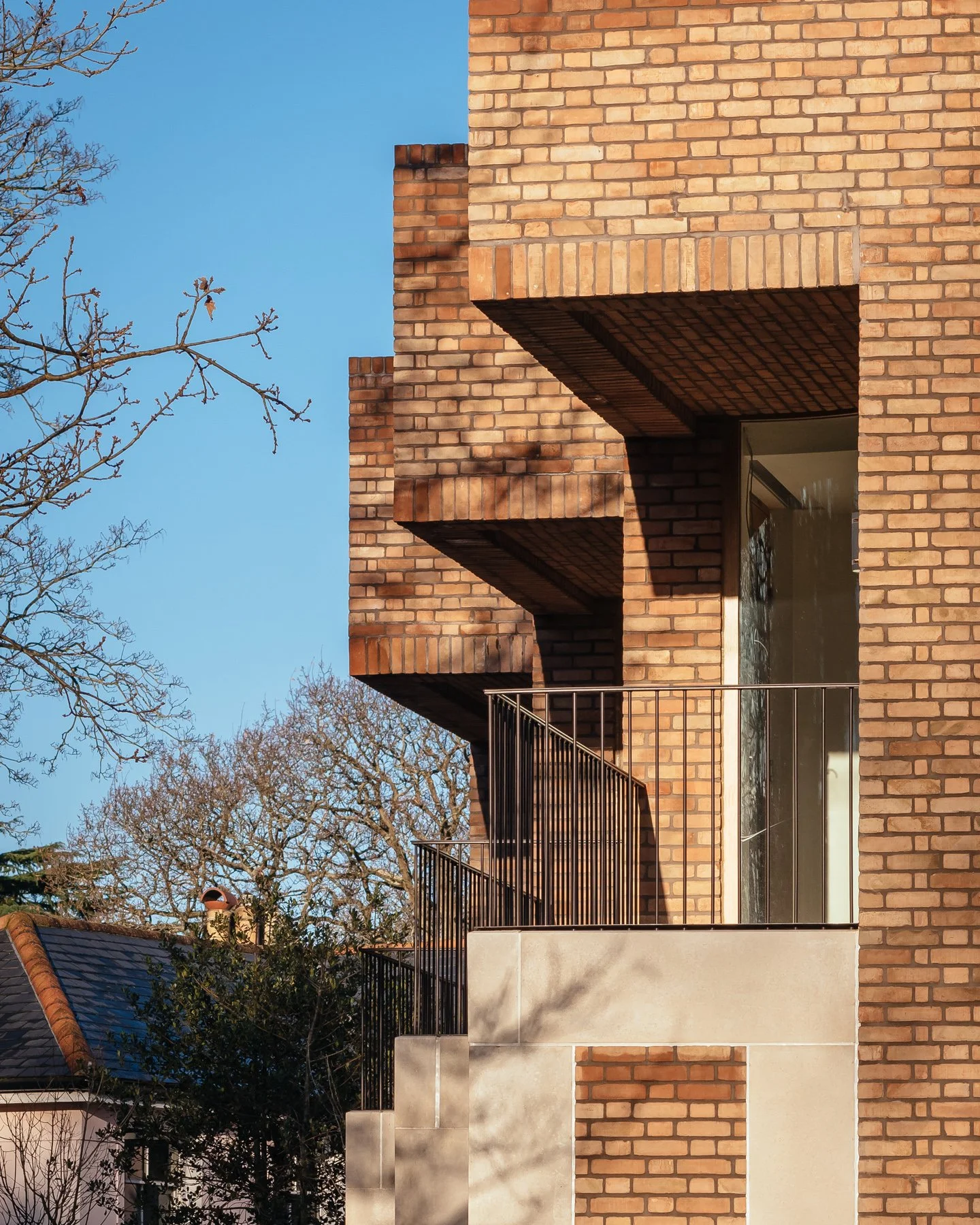 Close-up of a modern brick apartment building with balconies, large glass windows, and a metal railing, with leafless trees in the foreground and a clear blue sky in the background.