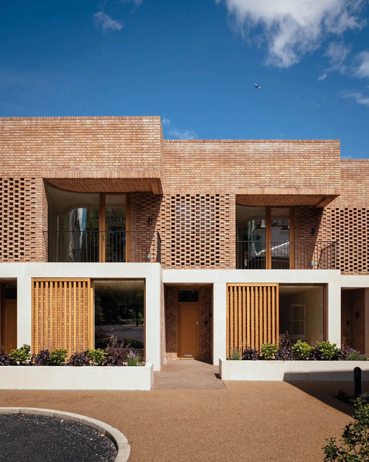 Modern brick townhouse with white accents, wood doors, and flower beds in front, under a blue sky.