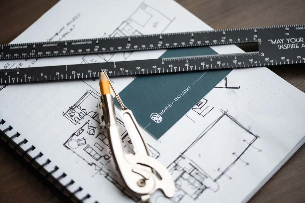 Blueprints, a ruler, and a drafting compass on a table, with a dark blue card labeled 'House of Daylight' resting on top of the blueprints.