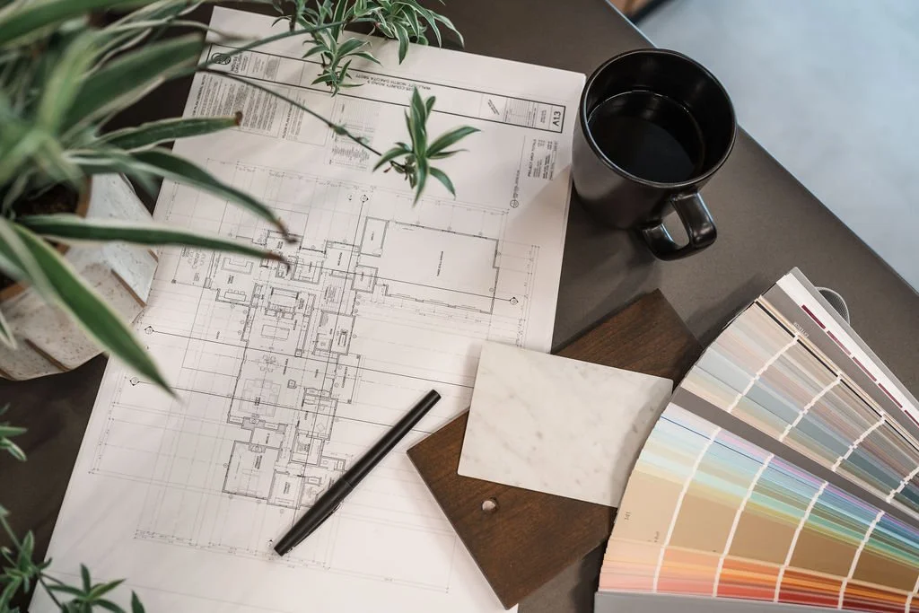 Architectural blueprint on desk with black coffee mug, color swatch samples, marble tile sample, and potted plants.
