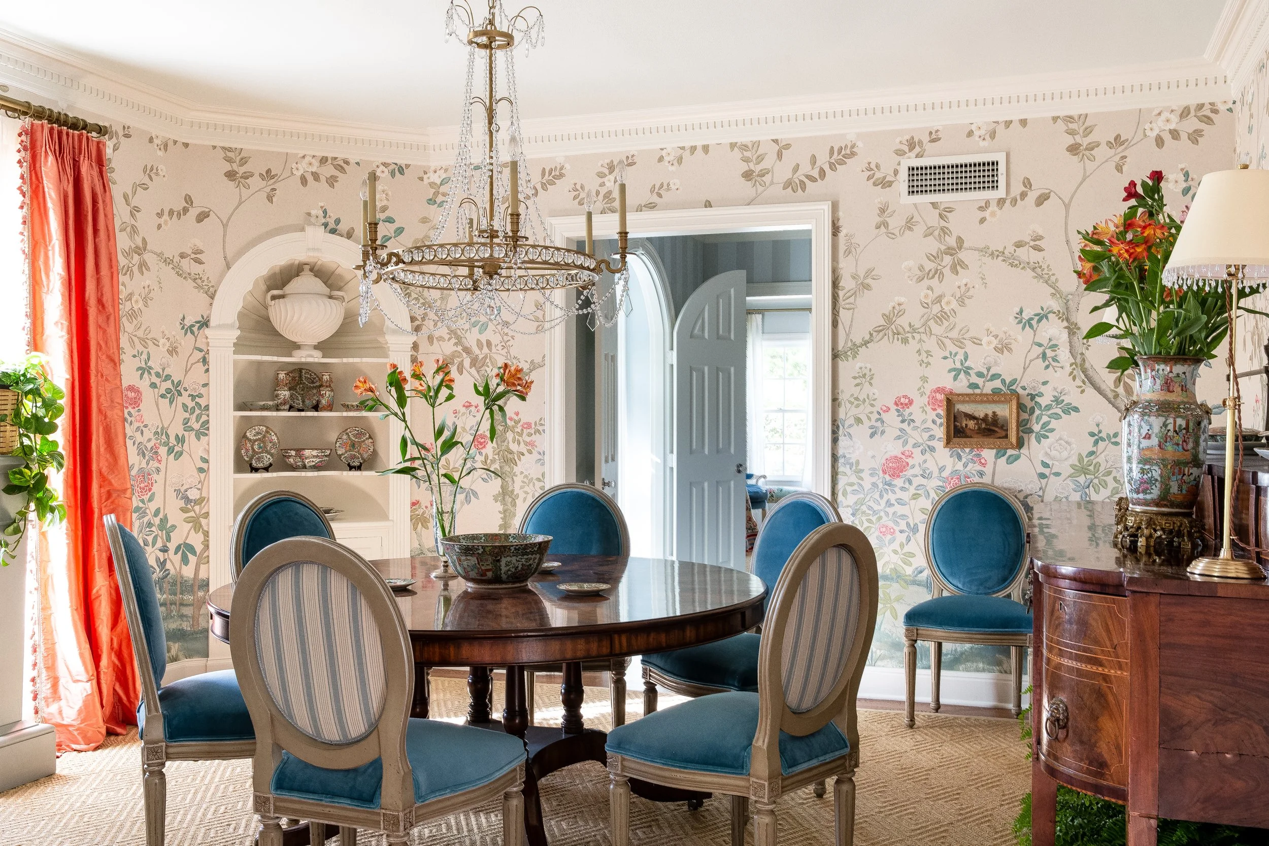 An elegant dining room featuring a round wooden table surrounded by blue velvet chairs, with floral wallpaper, a crystal chandelier, and lush greenery adding a touch of sophistication by Mark D. Sikes