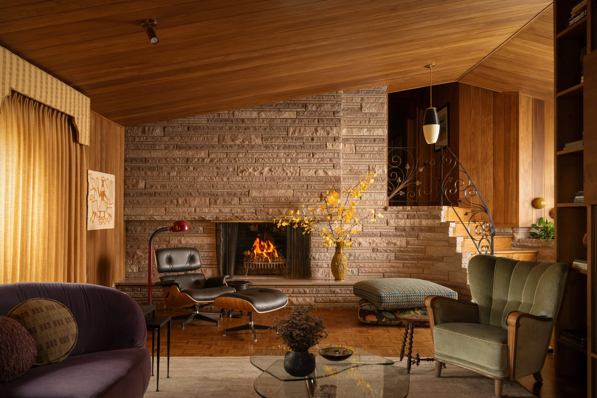 Living room with brick fireplace and wooden walls, vintage furniture including a green armchair and a black lounge chair, a glass coffee table with a vase and bowl, a yellow floral arrangement, and a staircase with ornate iron railing. Captured by Je