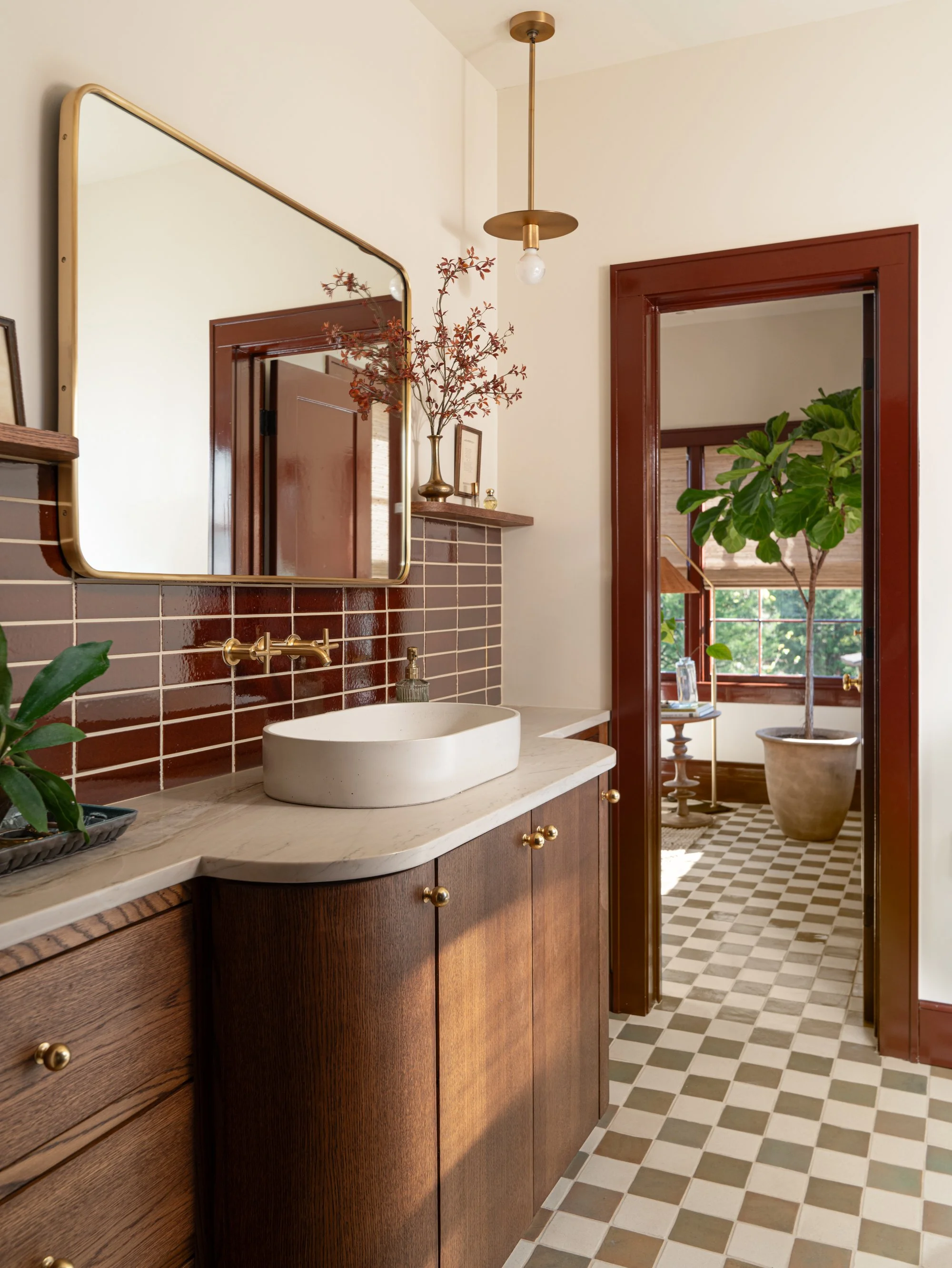 A bathroom with a wooden vanity, a white vessel sink, a large mirror, and a patterned tile floor. There are plants and decorative items, with a doorway leading to a room with a large potted tree and windows. Captured by Jeff Jones of Jeff Jones Photo