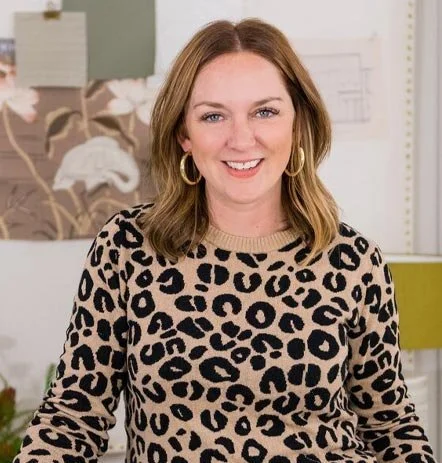 Kate Bendewald. A woman with shoulder-length hair, wearing hoop earrings and a leopard print sweater, smiling in an indoor setting.