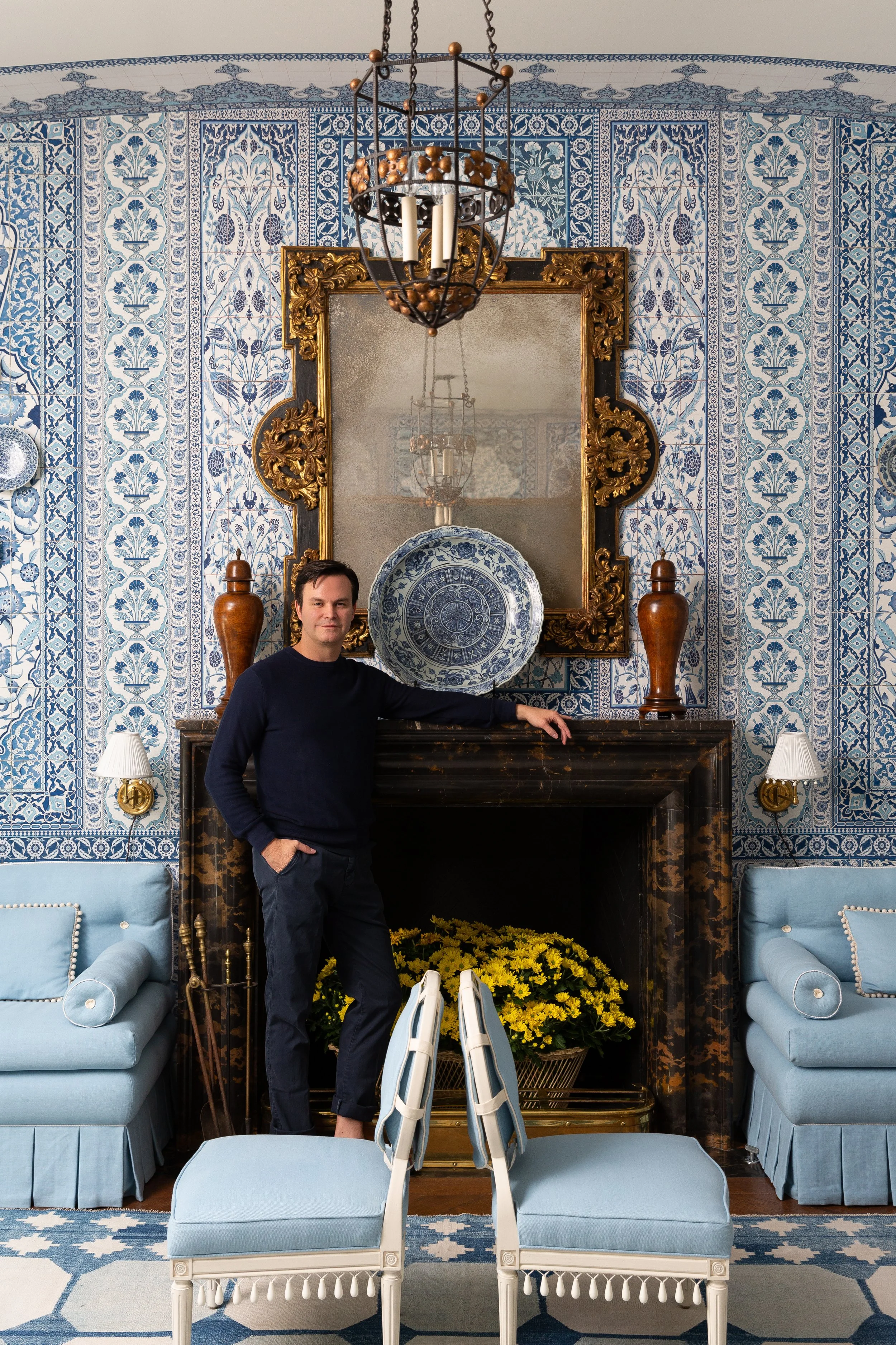 A man in a black sweater and dark pants standing in their living room with one hand on the black marble fireplace mantel, surrounded by light blue furniture, yellow flowers, and blue patterned wallpaper.
