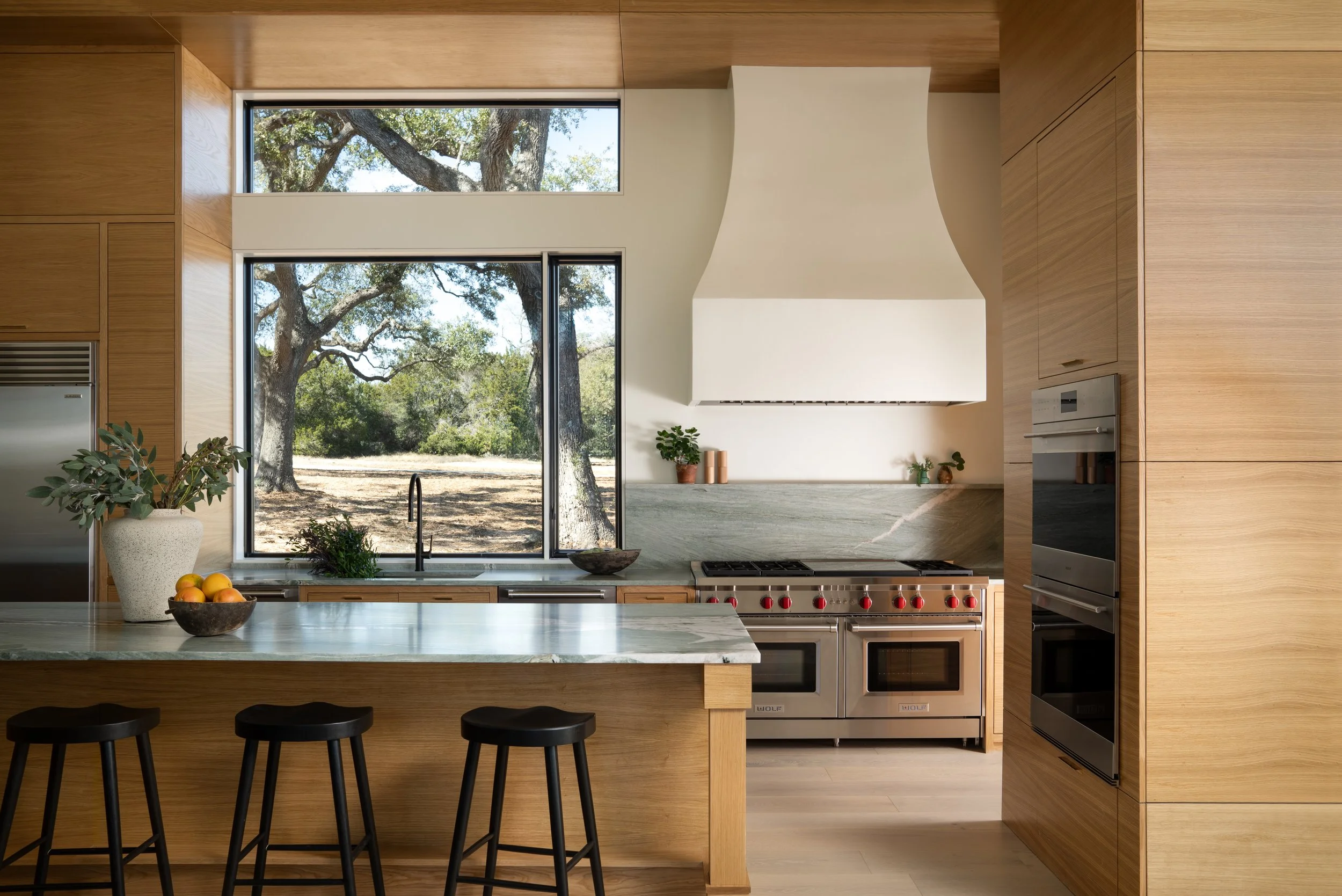 Interior design photography by Jeff Jones featuring a modern kitchen with wood paneling, Wolf stove, green marble countertops, and a large picture window. Showcasing asymmetry design in Austin, TX.